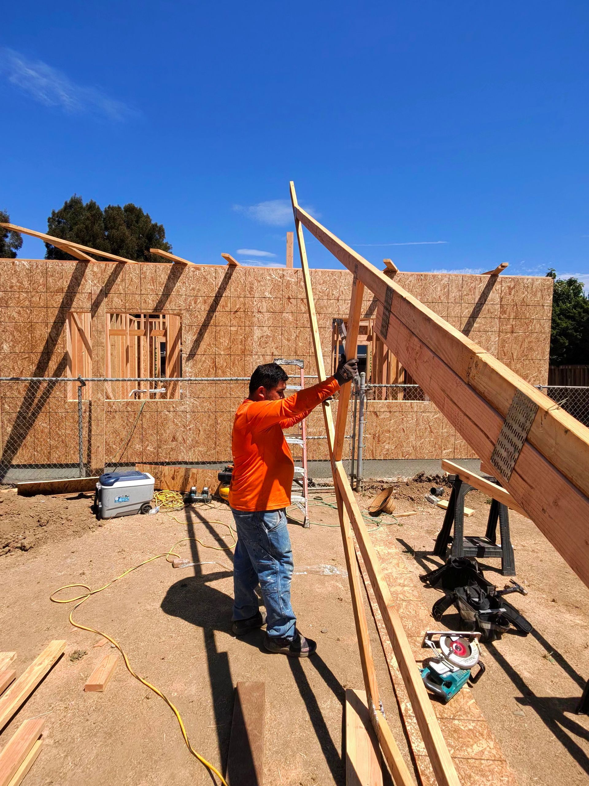 Worker in orange shirt raising a roof truss at a house construction site under a clear blue sky