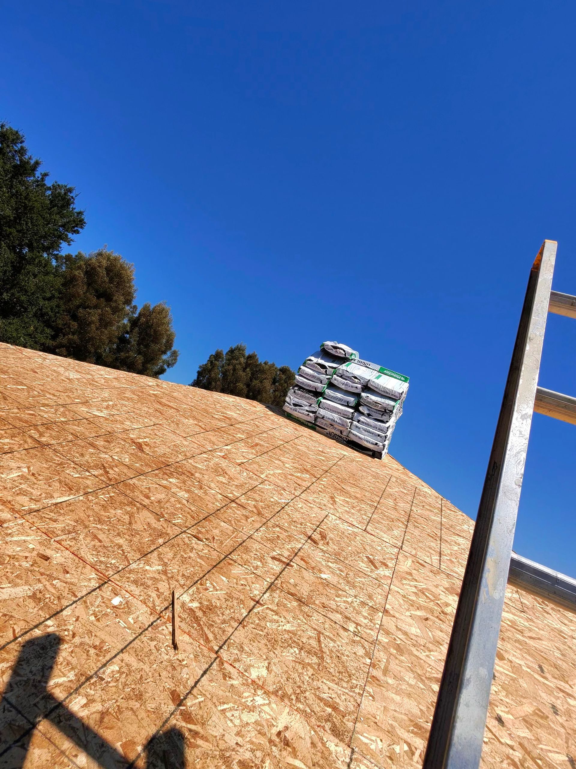 Tilted view of a rooftop with a ladder, a vent, and trees against a clear blue sky