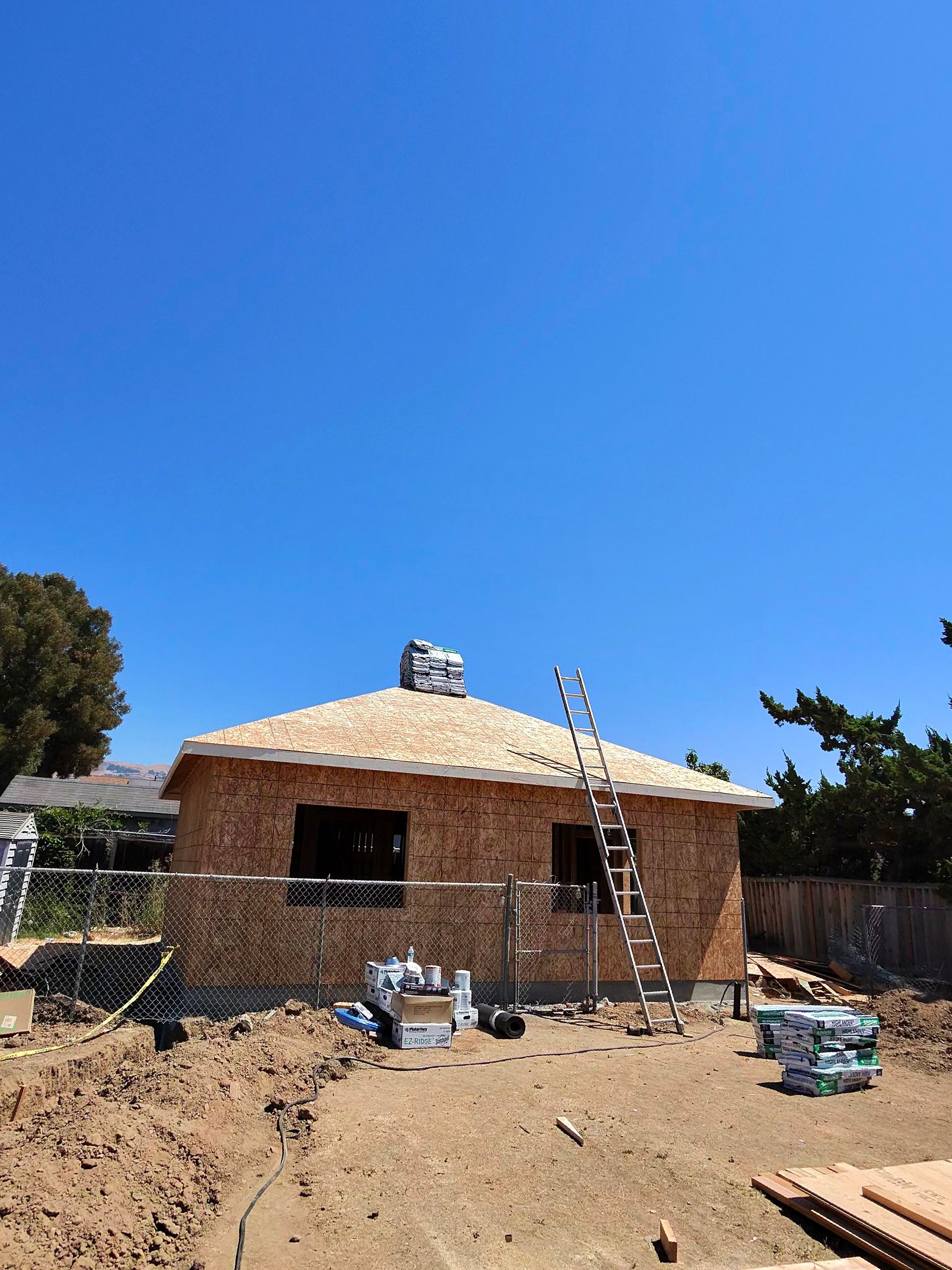 Partially built brick house under construction with scaffolding and building materials in a dirt yard under a blue sky