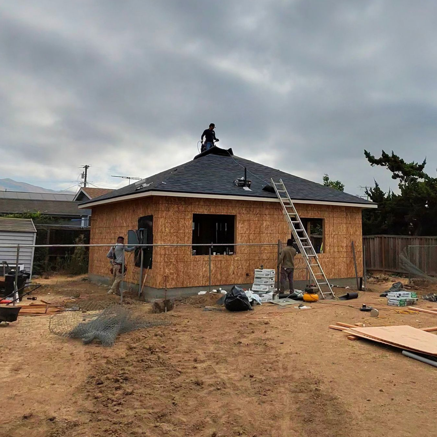 Single-story house under construction with a worker on the roof and ladders in a dirt yard