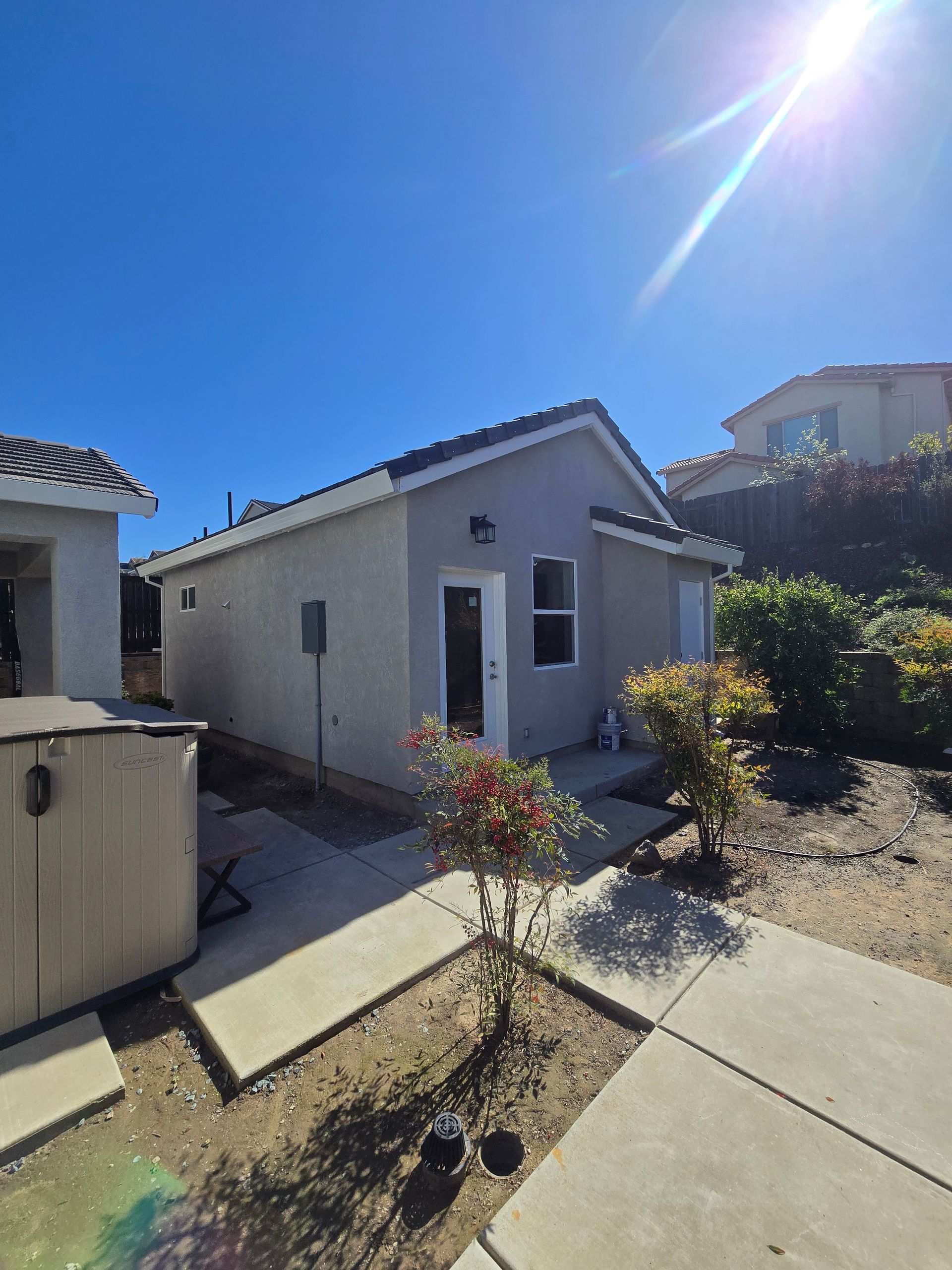 Small stucco house with a tiled roof beside a concrete path under bright sunlight.