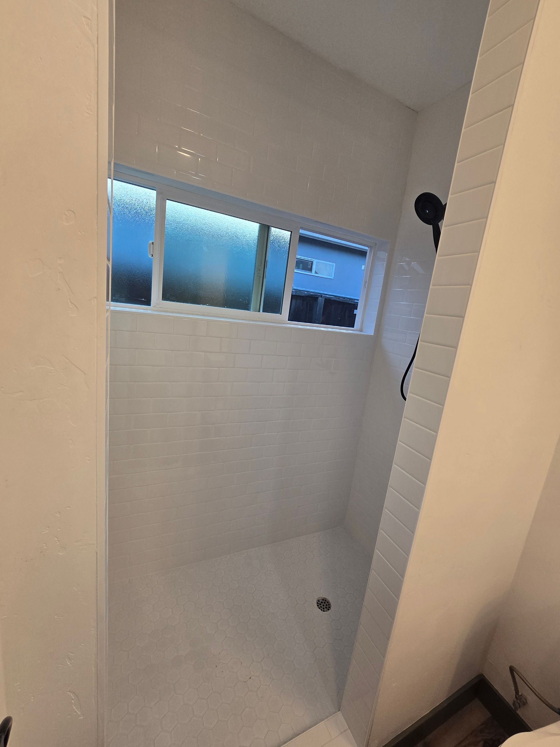 Empty white tiled shower stall with a small window near the top and a drain in the floor.