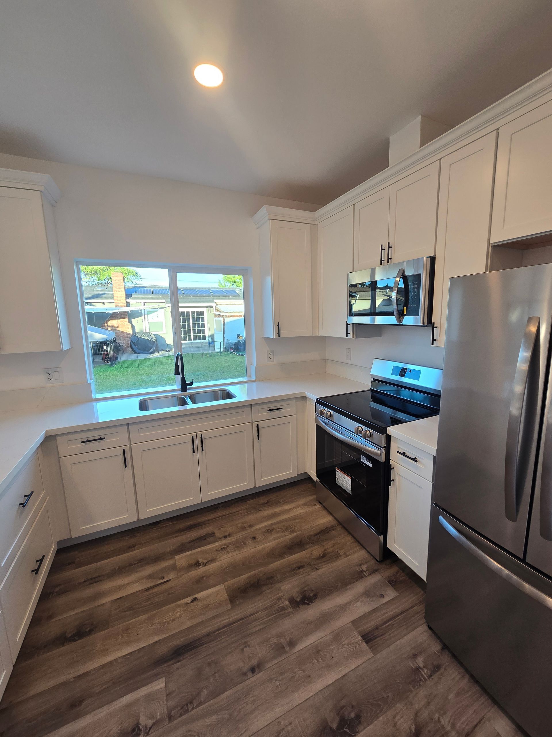 Bright white kitchen with stainless steel appliances, wood-look floors, and a window overlooking a backyard.