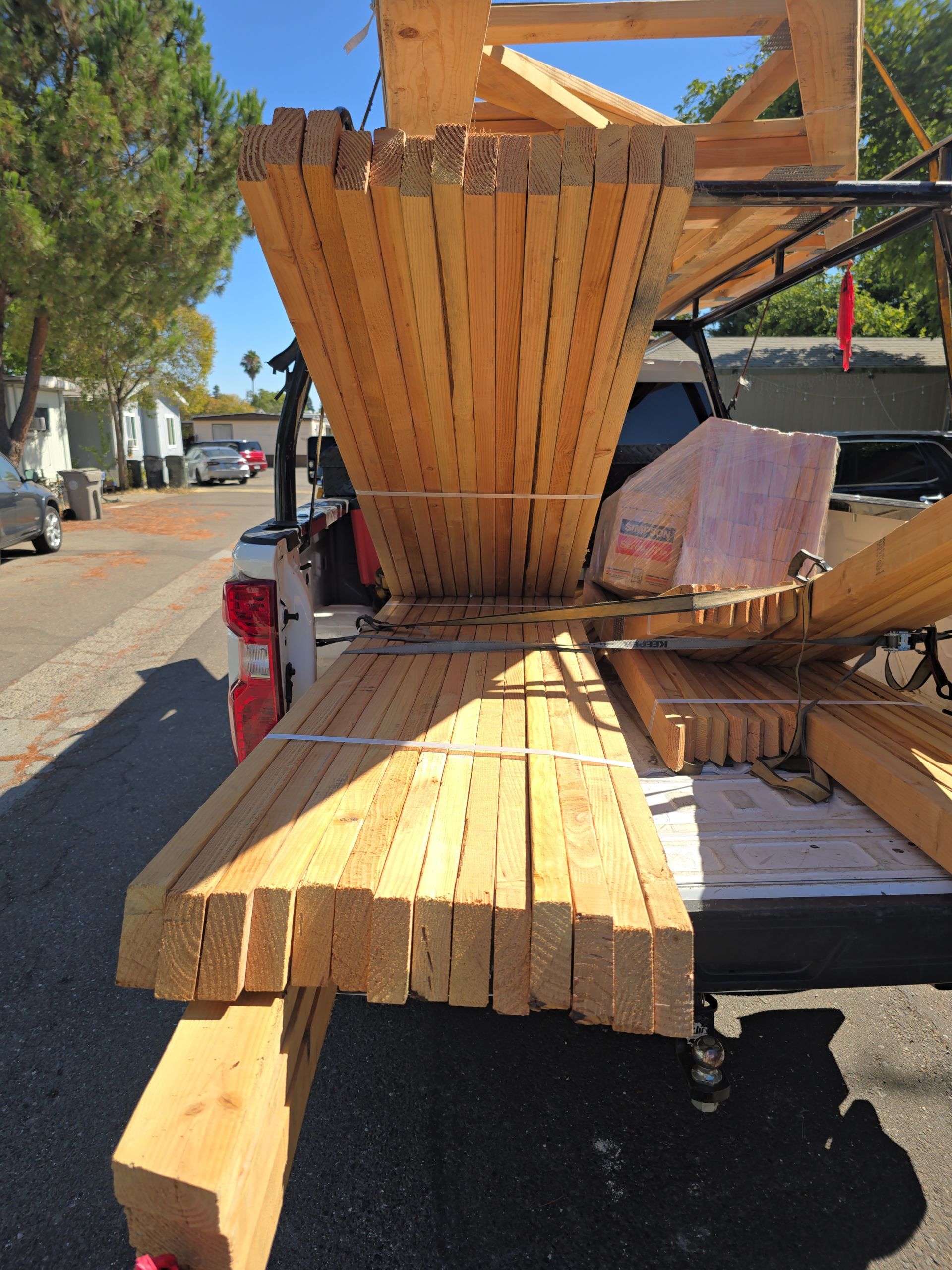 Pickup truck bed loaded with long wooden boards and lumber, parked outdoors on a sunny street.