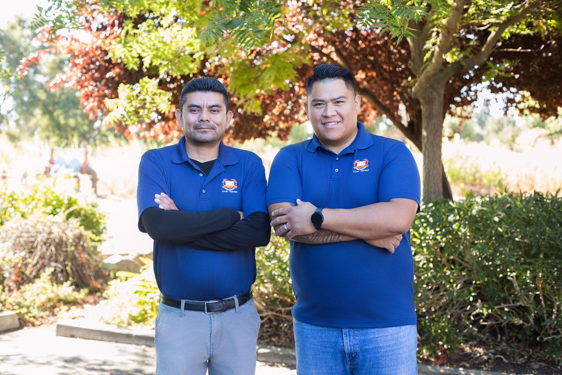 Two men in blue polo shirts stand with arms crossed in a sunny garden.