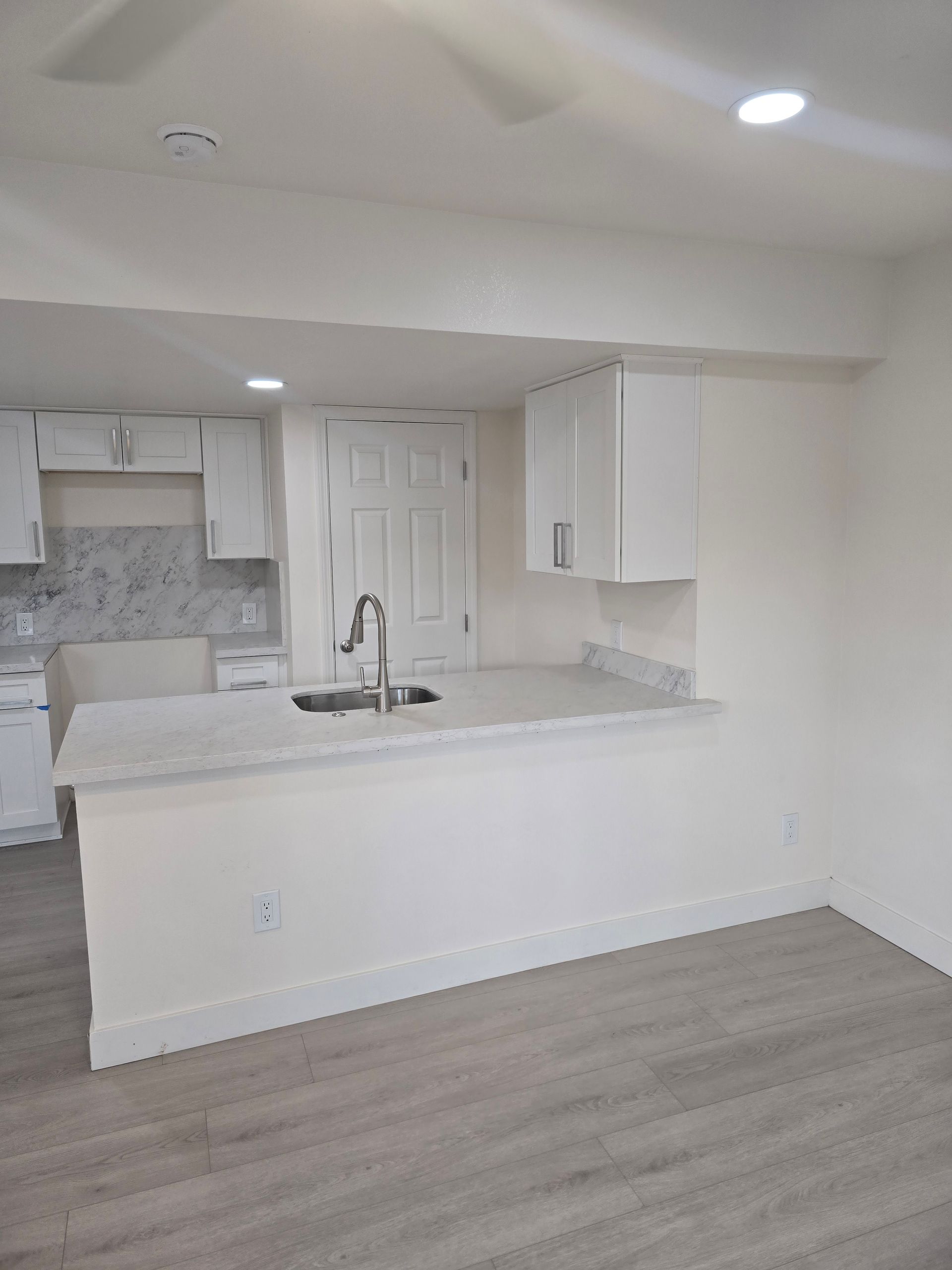 Bright white kitchen with island sink, cabinets, and light wood flooring in an empty room