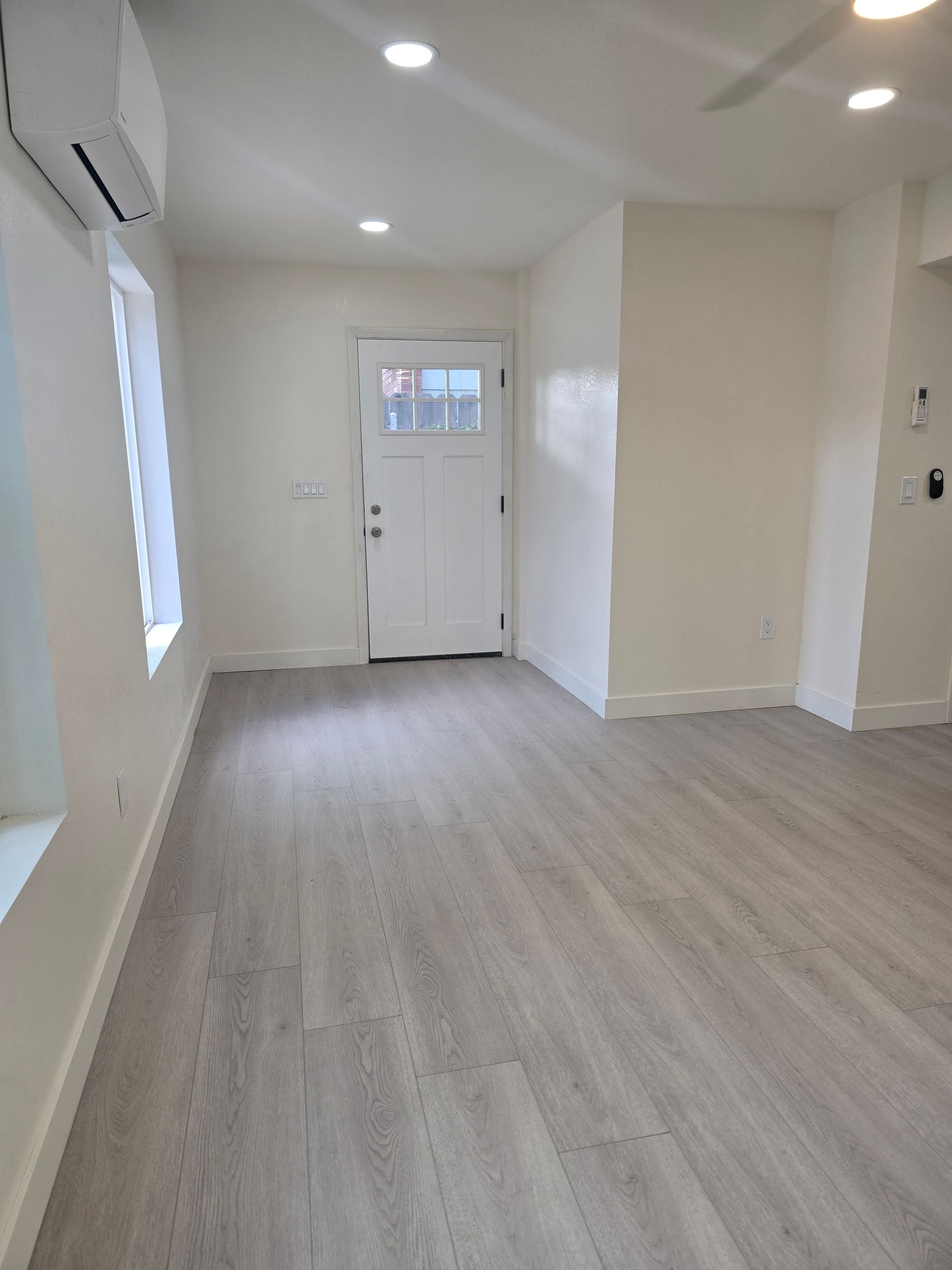 Empty modern room with light wood floor, white walls, recessed lights, and a front door with window.