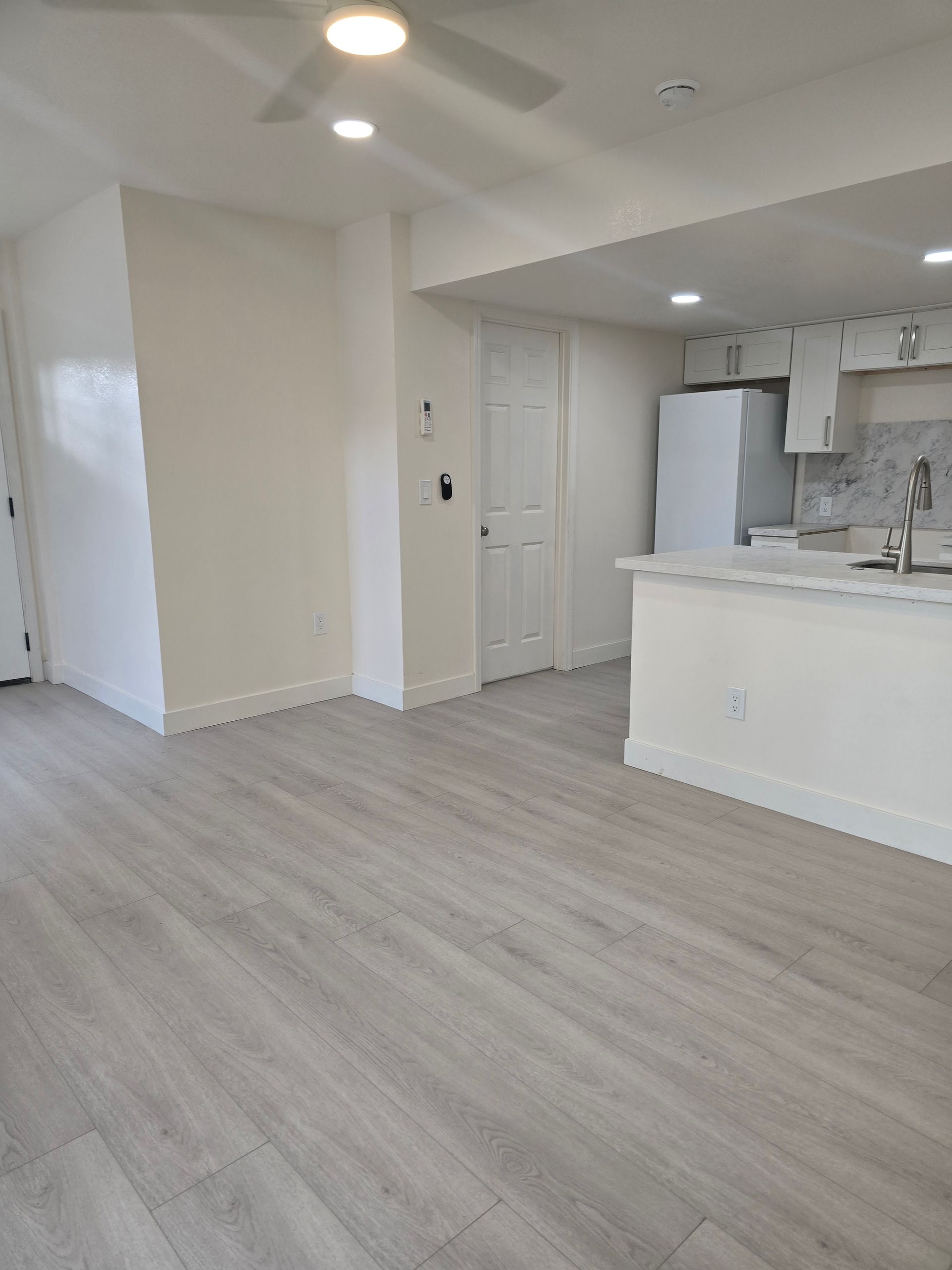 Empty modern kitchen and living area with light wood floors, white walls, and recessed ceiling lights