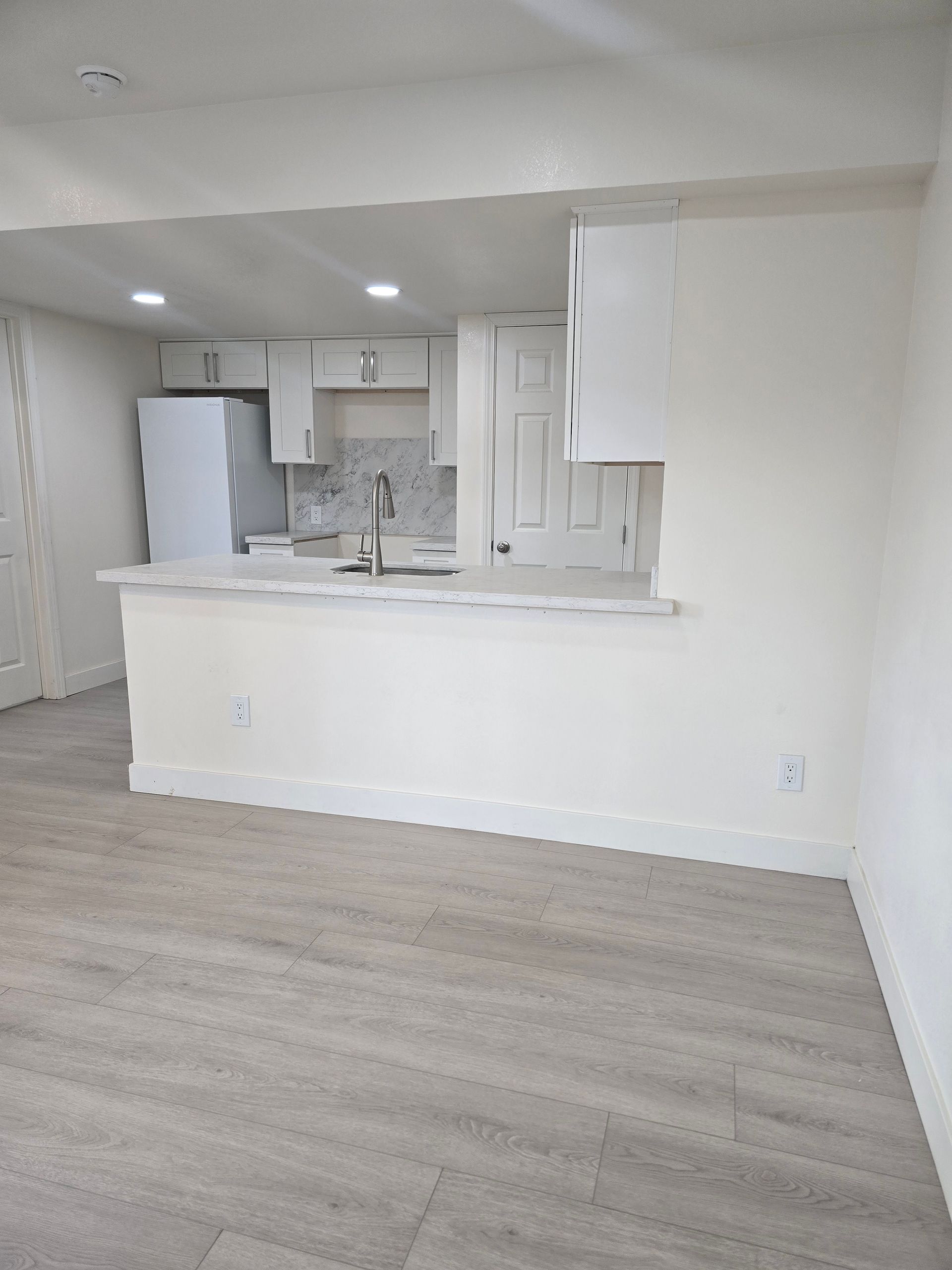 Bright, empty open-plan room with white kitchen pass-through and light gray wood flooring