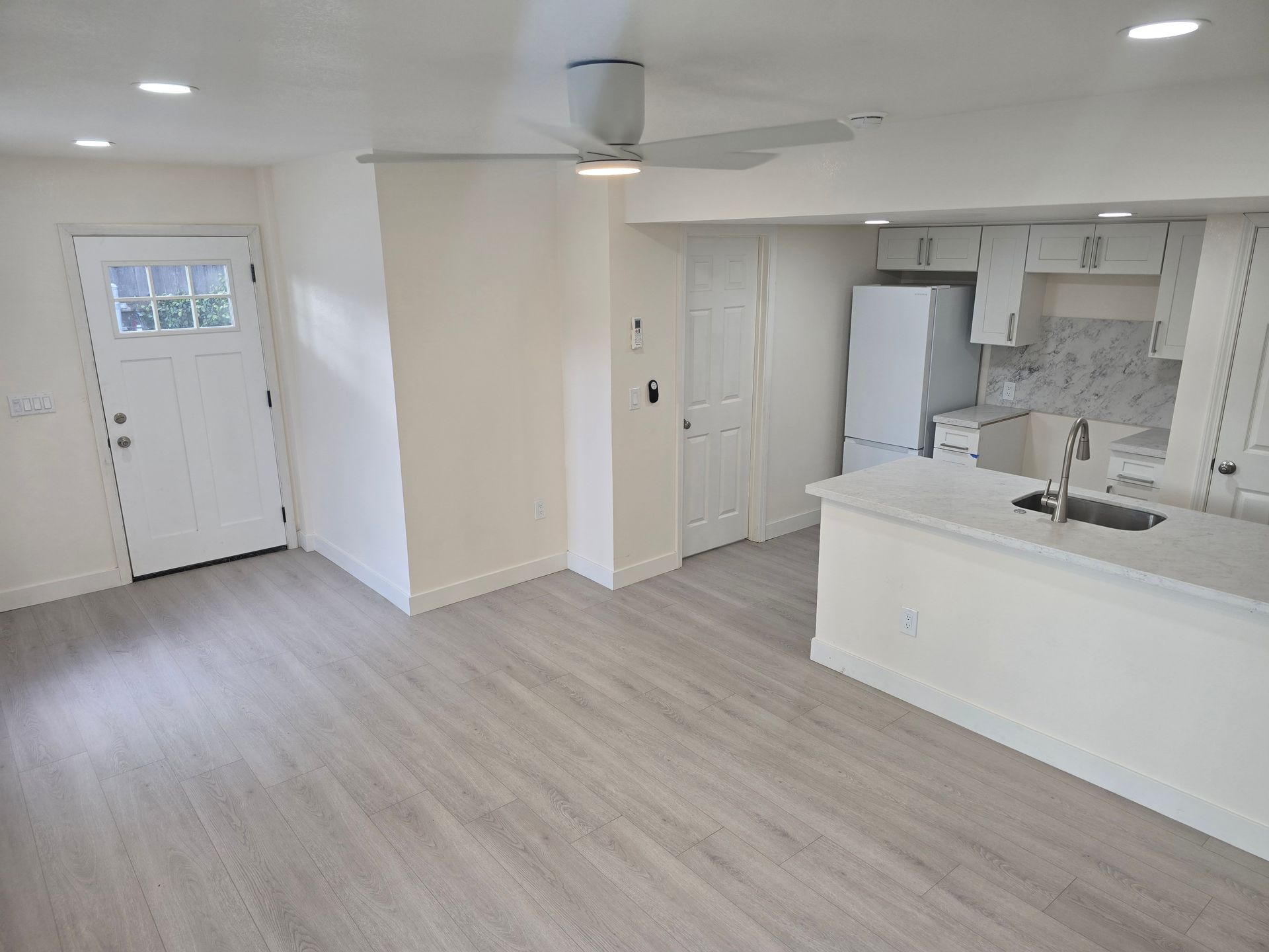 Bright empty kitchen and living area with white cabinets, light wood floors, and front door visible