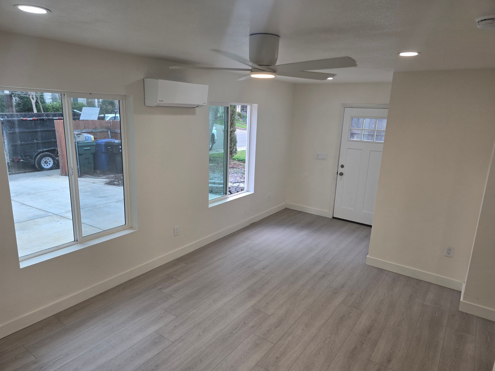 Empty beige living room with ceiling fan, windows, and front door with wood-look flooring