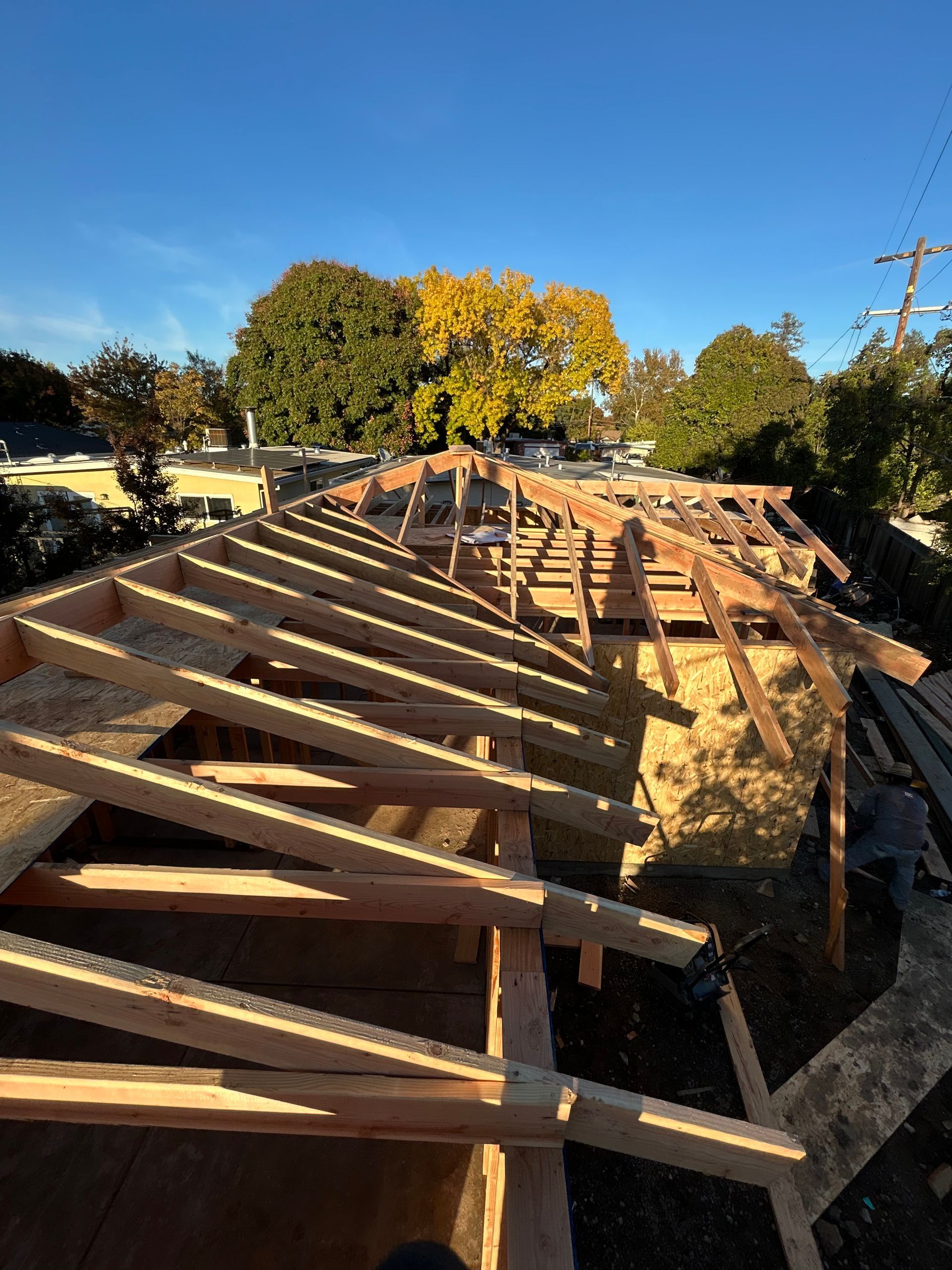 Roof framing under construction on a house, with exposed wooden beams and blue sky overhead