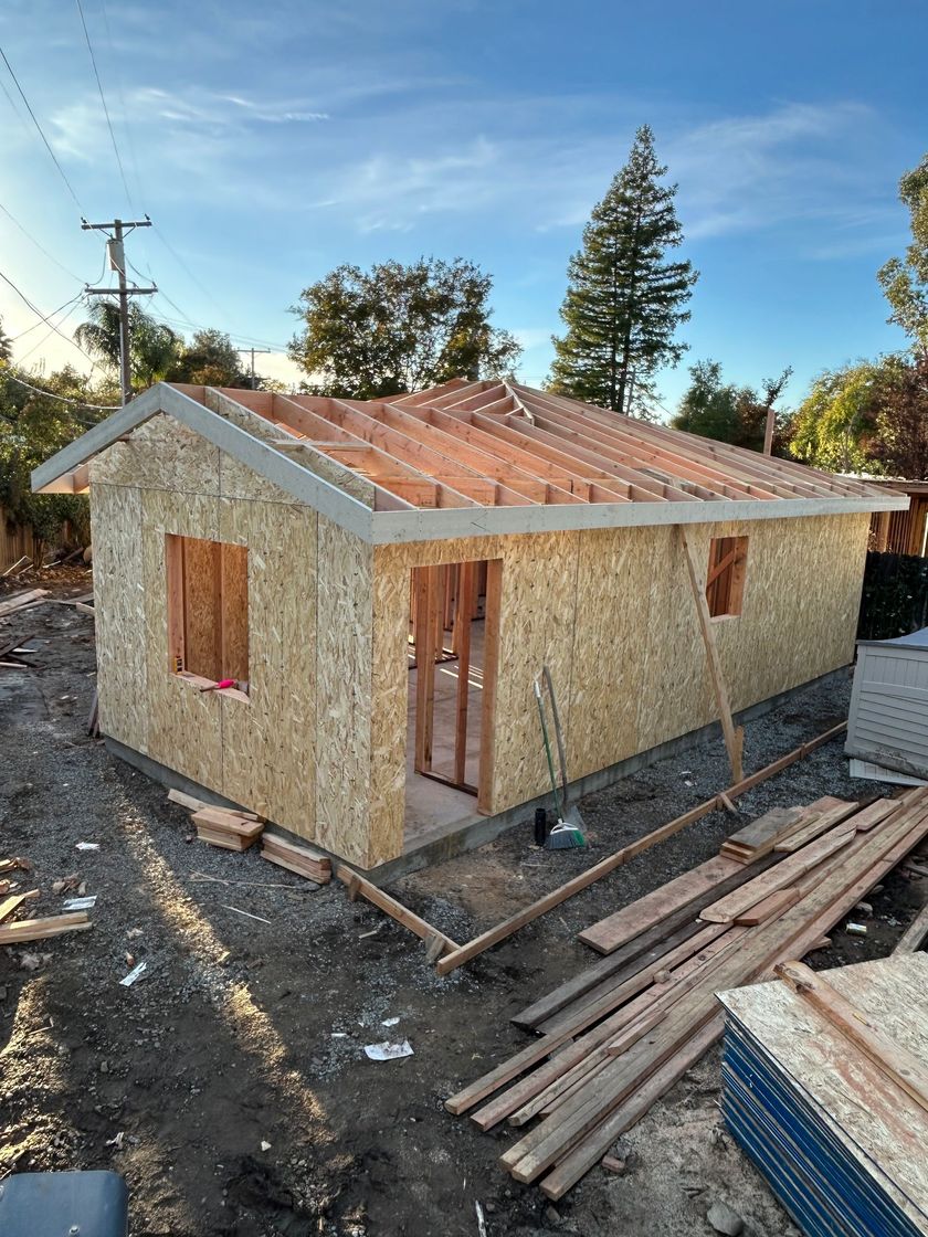 Small unfinished house with a rusty metal roof and boarded windows at a construction site.