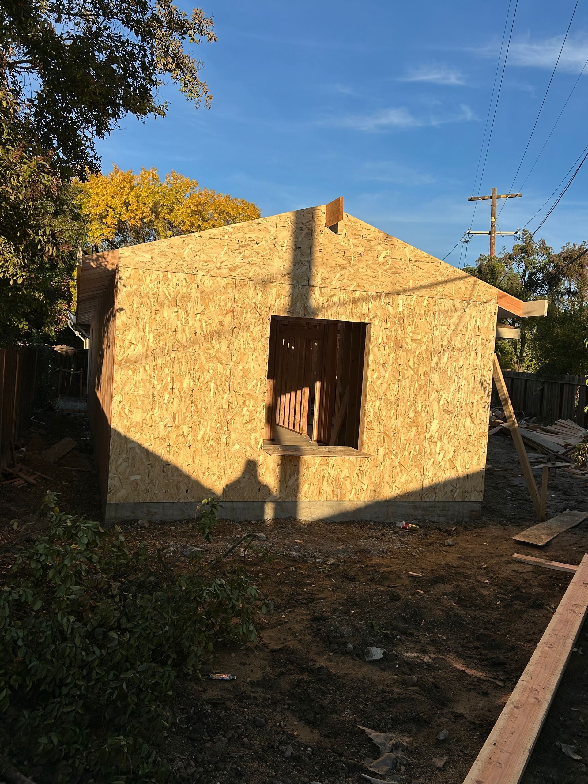 Small unfinished brick house with a window, lit by sunlight in a dirt yard with trees nearby.
