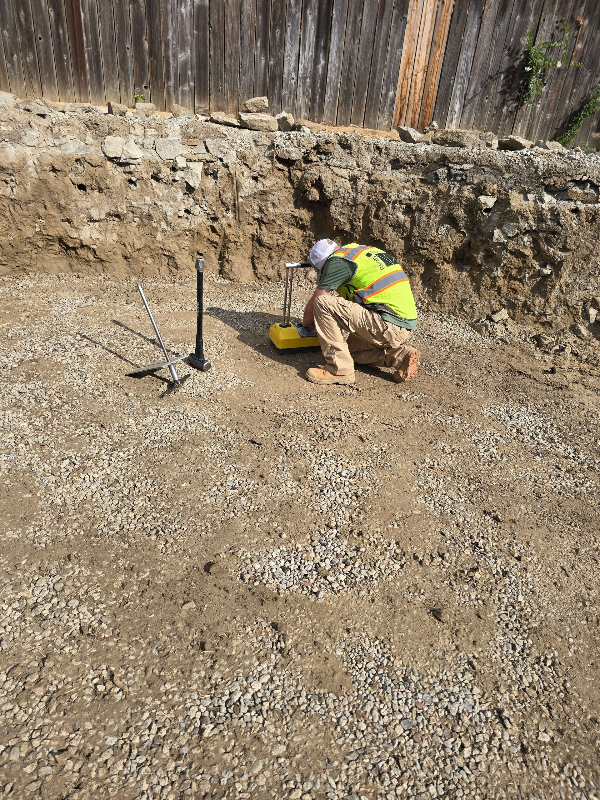 Worker in a neon safety vest kneels in a dirt excavation site, examining a yellow bucket near a shovel.