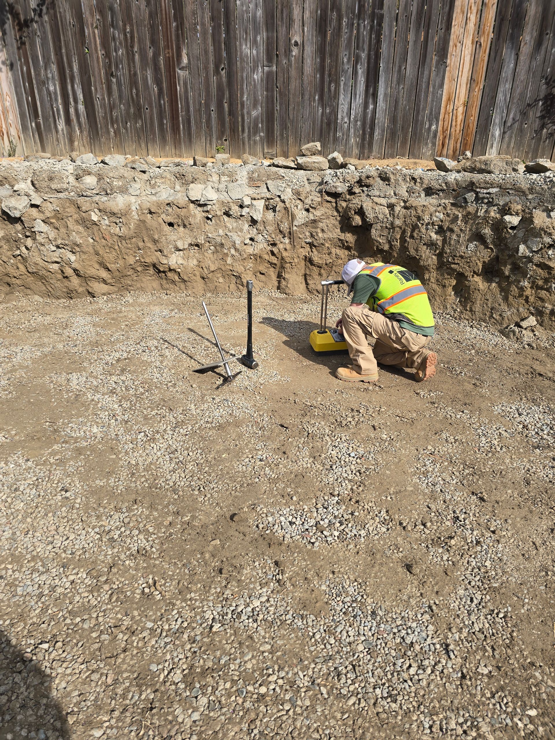 Construction worker in neon vest kneeling by a measuring tool in a dirt excavation site