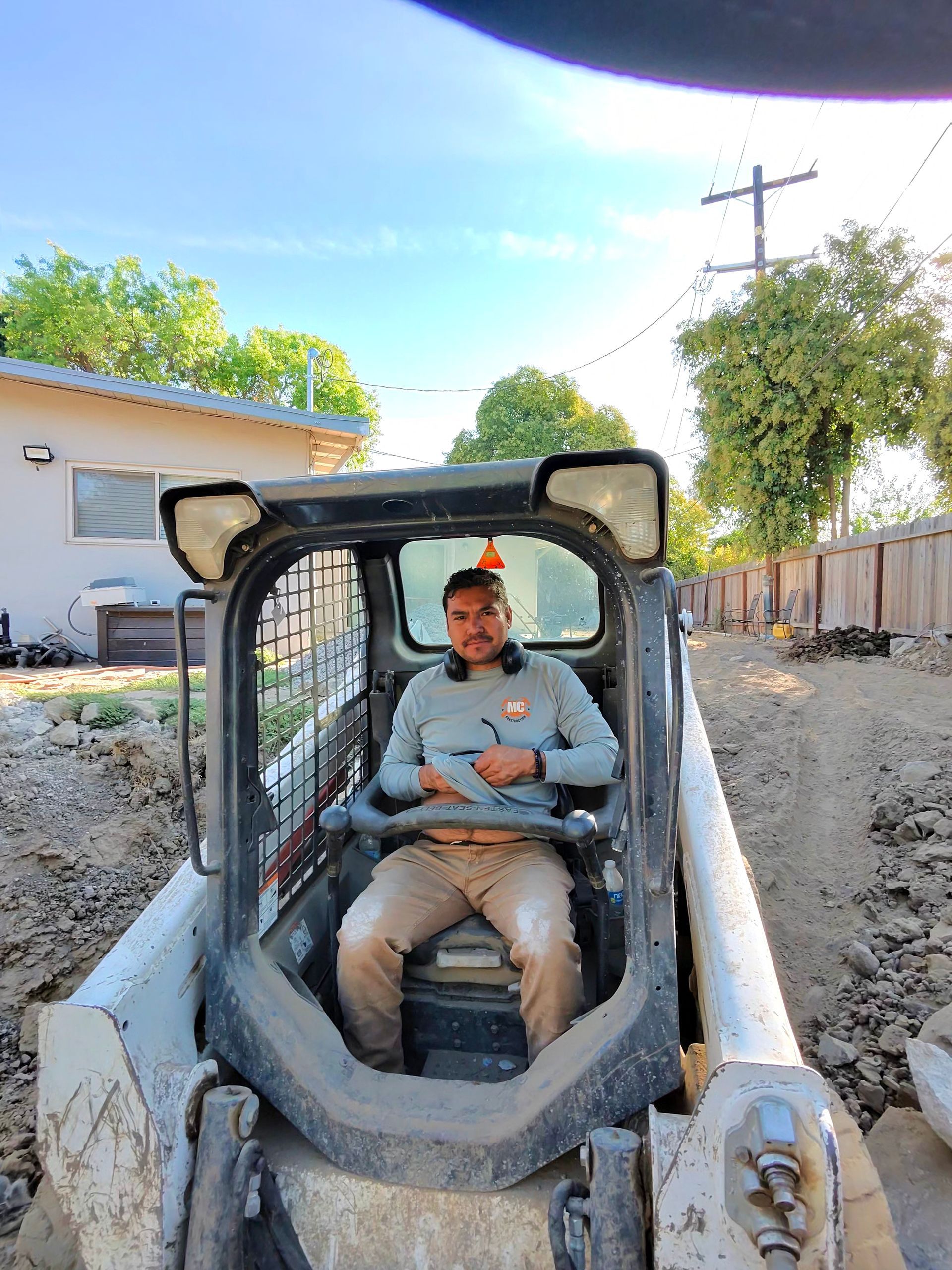 Worker seated in a compact excavator at a construction site, with dirt piles and fencing nearby.
