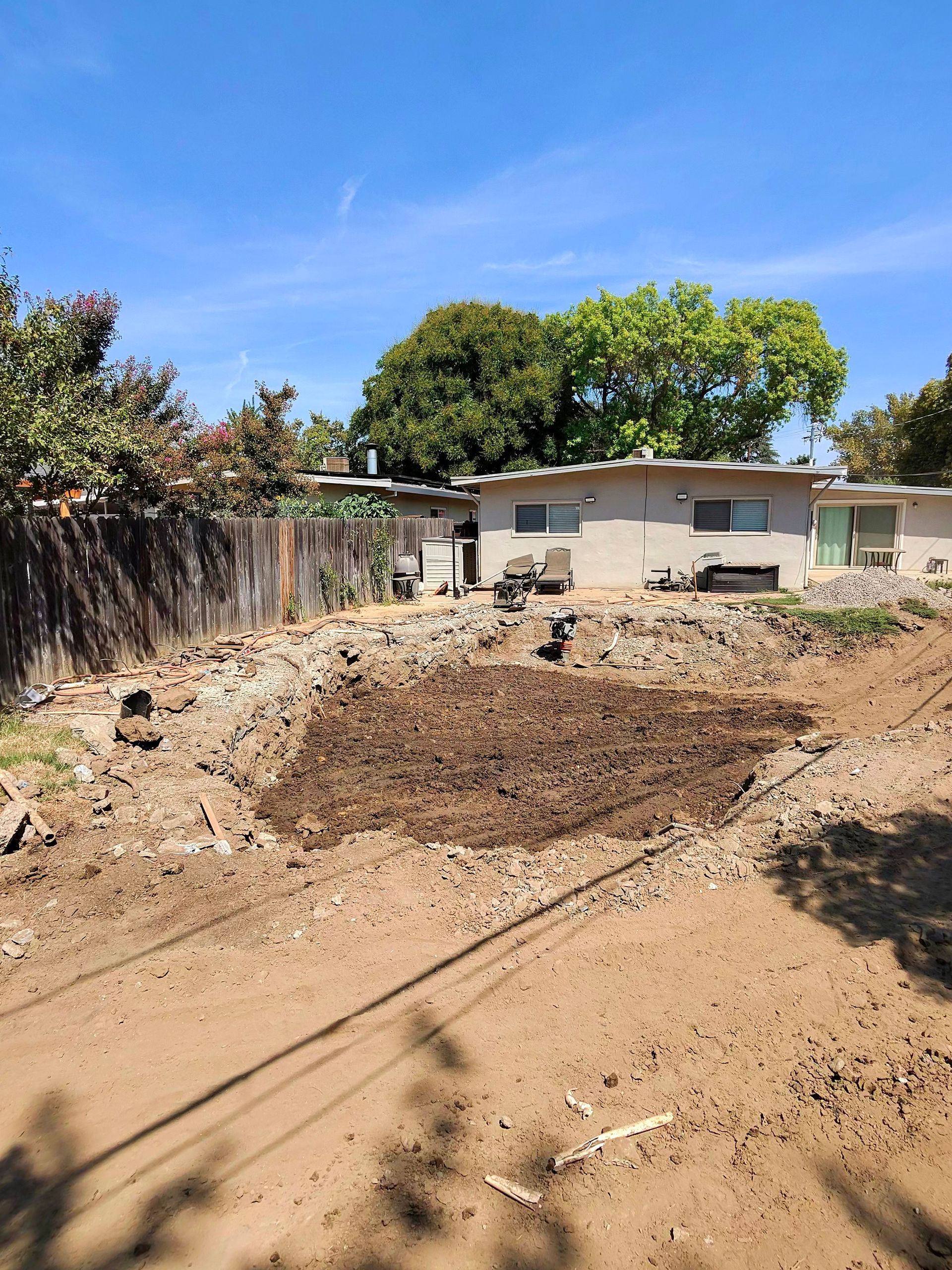 Backyard with a large dug-out dirt pit, fence, and a house under a blue sky