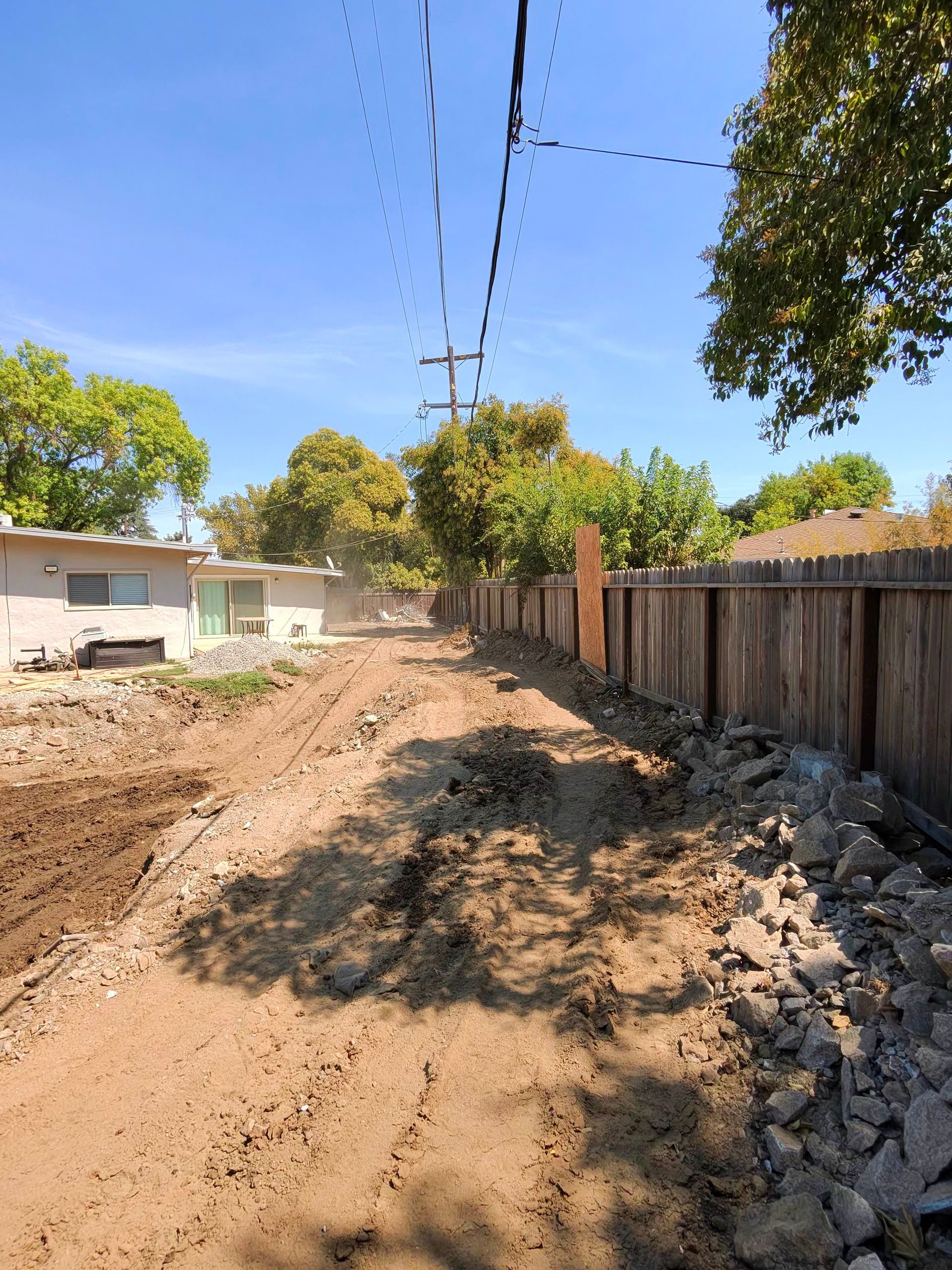 Dirt road under construction beside a wooden fence, with power lines overhead and trees lining the street.