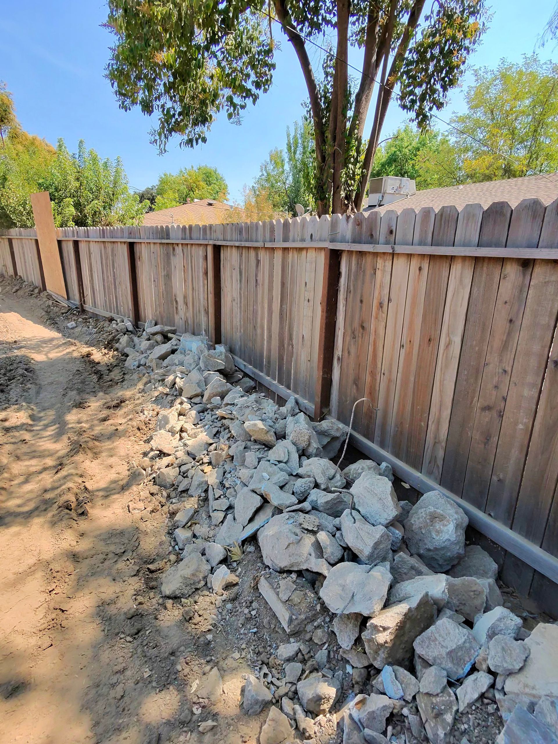 Rubble piled along a wooden fence beside a dirt path under trees in a sunny yard