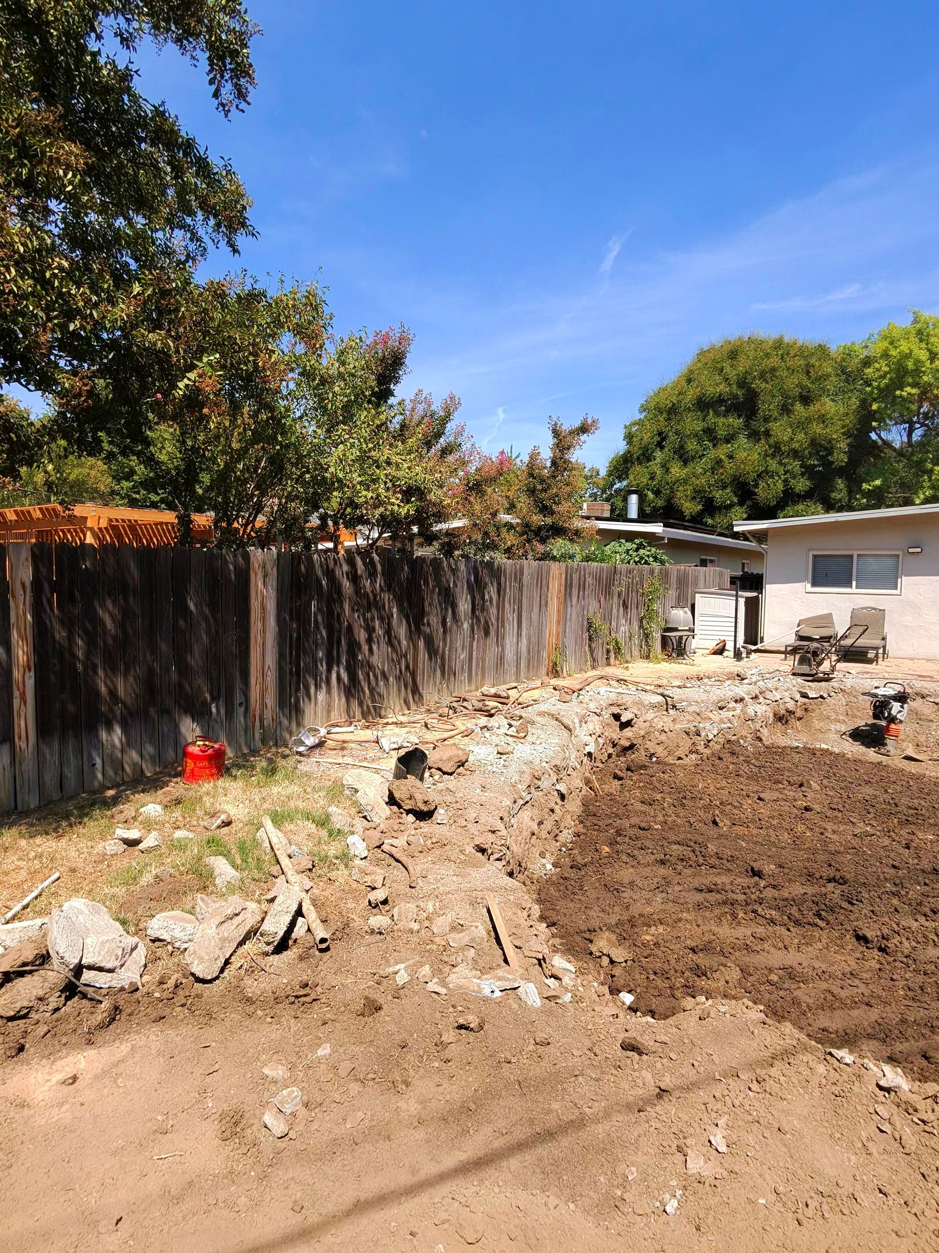 Mud-filled backyard beside a wooden fence, with trees, a small shed, and construction debris under a blue sky