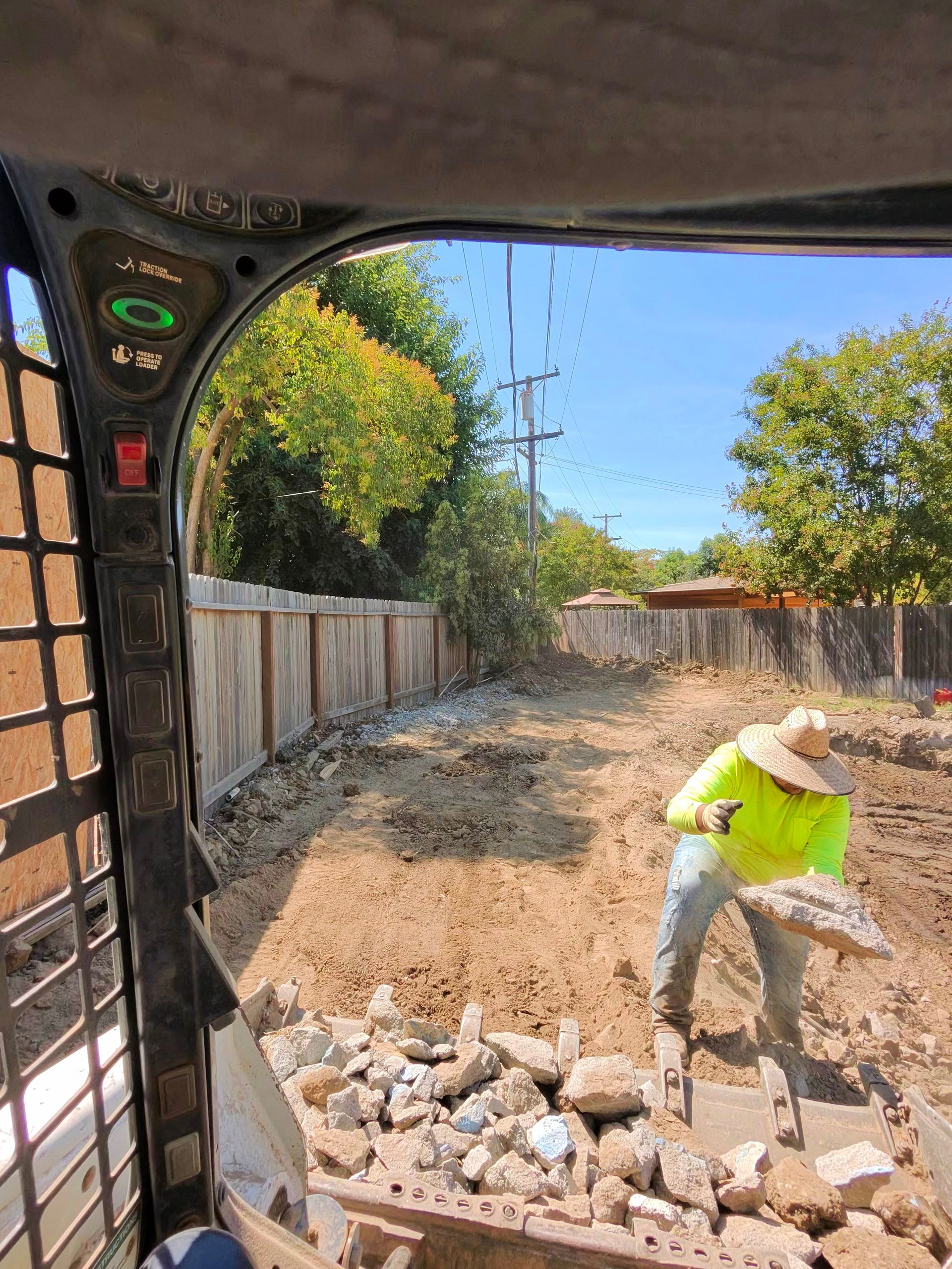 Construction worker breaking concrete in a backyard with rubble piles and a fence under a blue sky