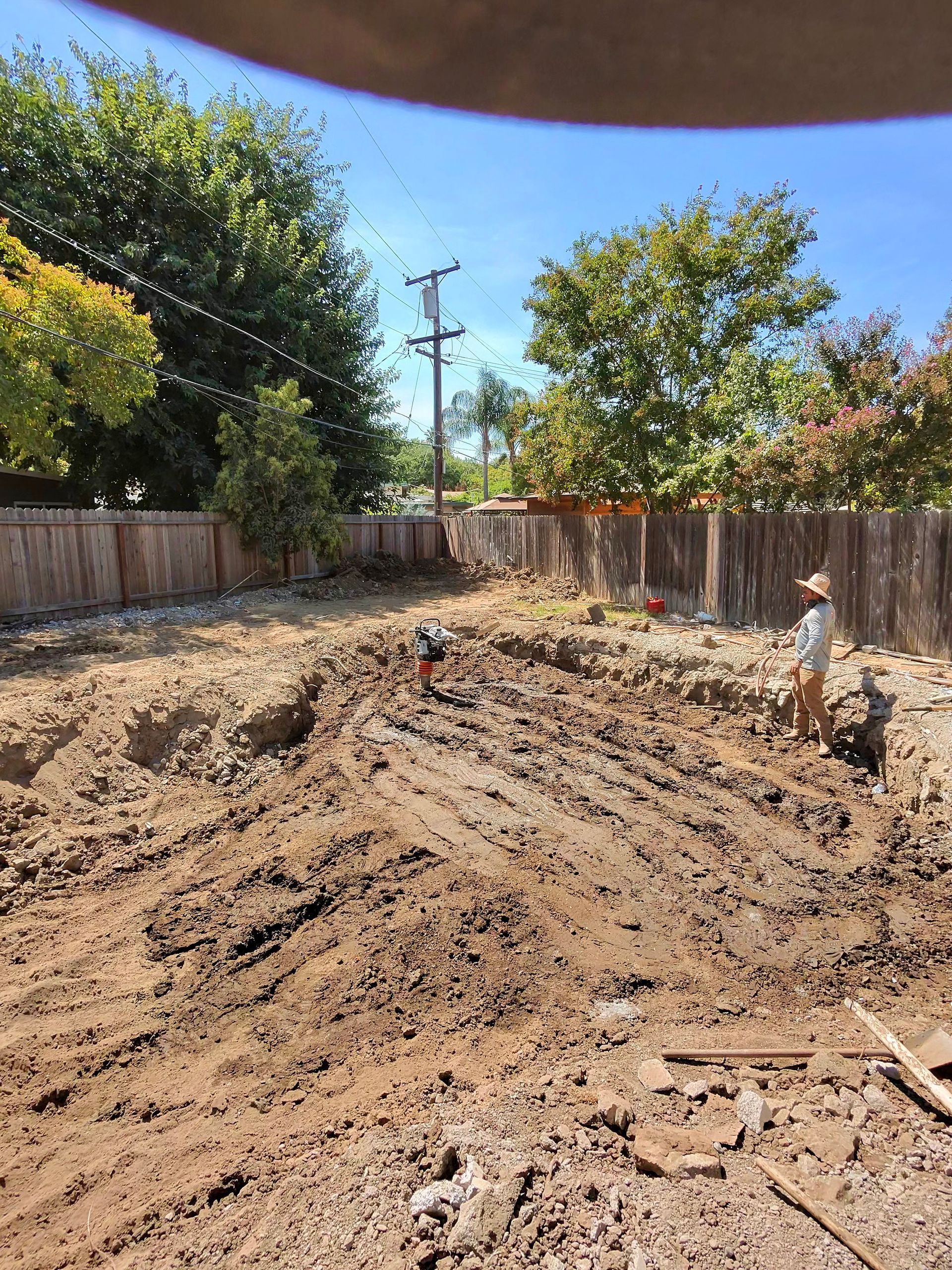 Backyard dirt mound with two workers and a shovel, surrounded by fences and trees on a sunny day