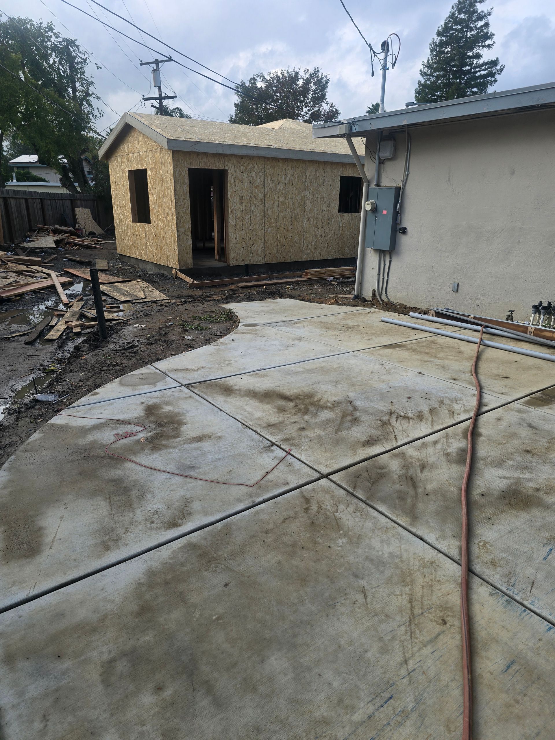 Concrete driveway beside a small unfinished shed and house wall, with debris and power lines overhead.