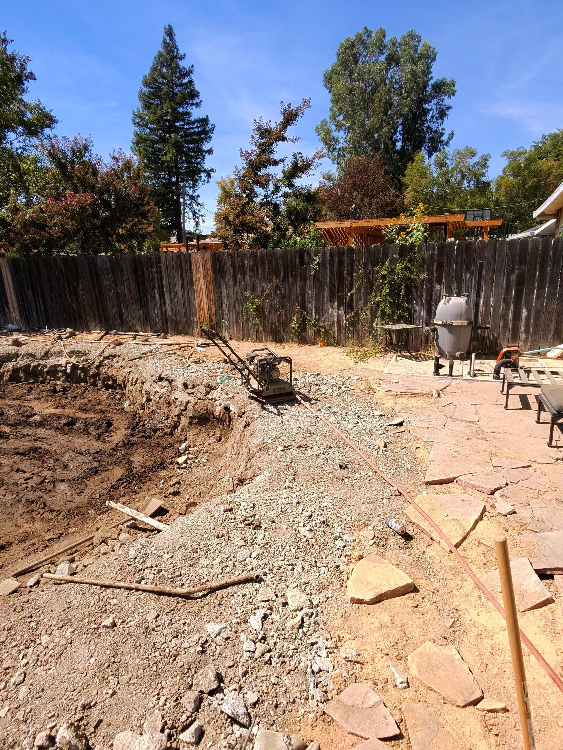 Backyard under construction with gravel, dirt, fence, and a wheelbarrow in the sun