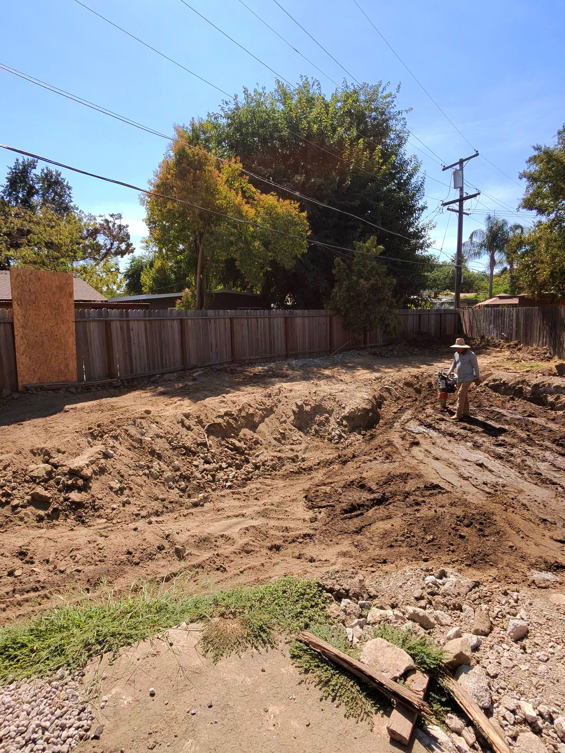 Dirt excavation site with a worker, wooden fence, trees, and utility poles under a clear blue sky
