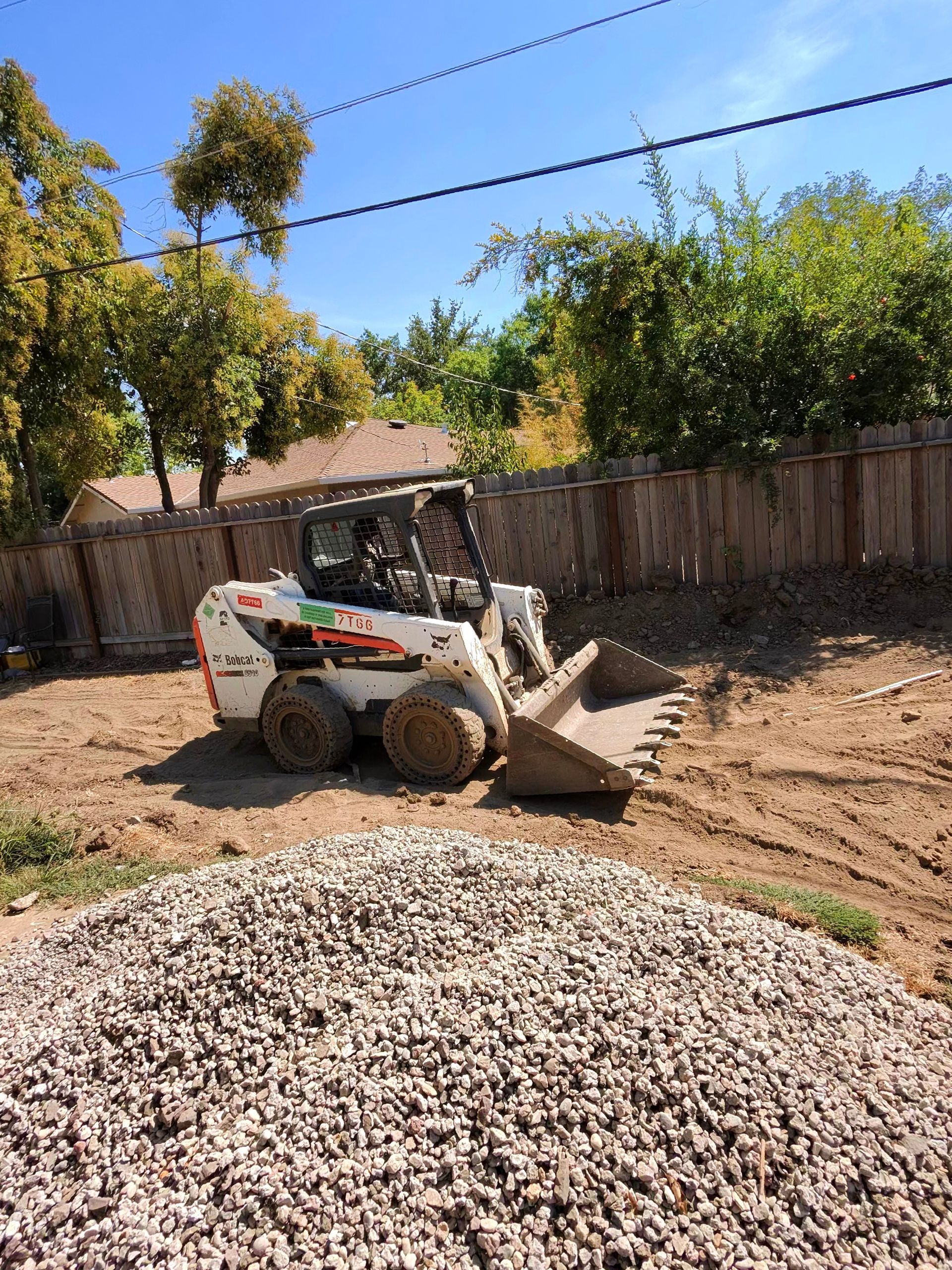 Small white skid-steer loader moving dirt in a dusty yard beside a gravel pile and wooden fence.