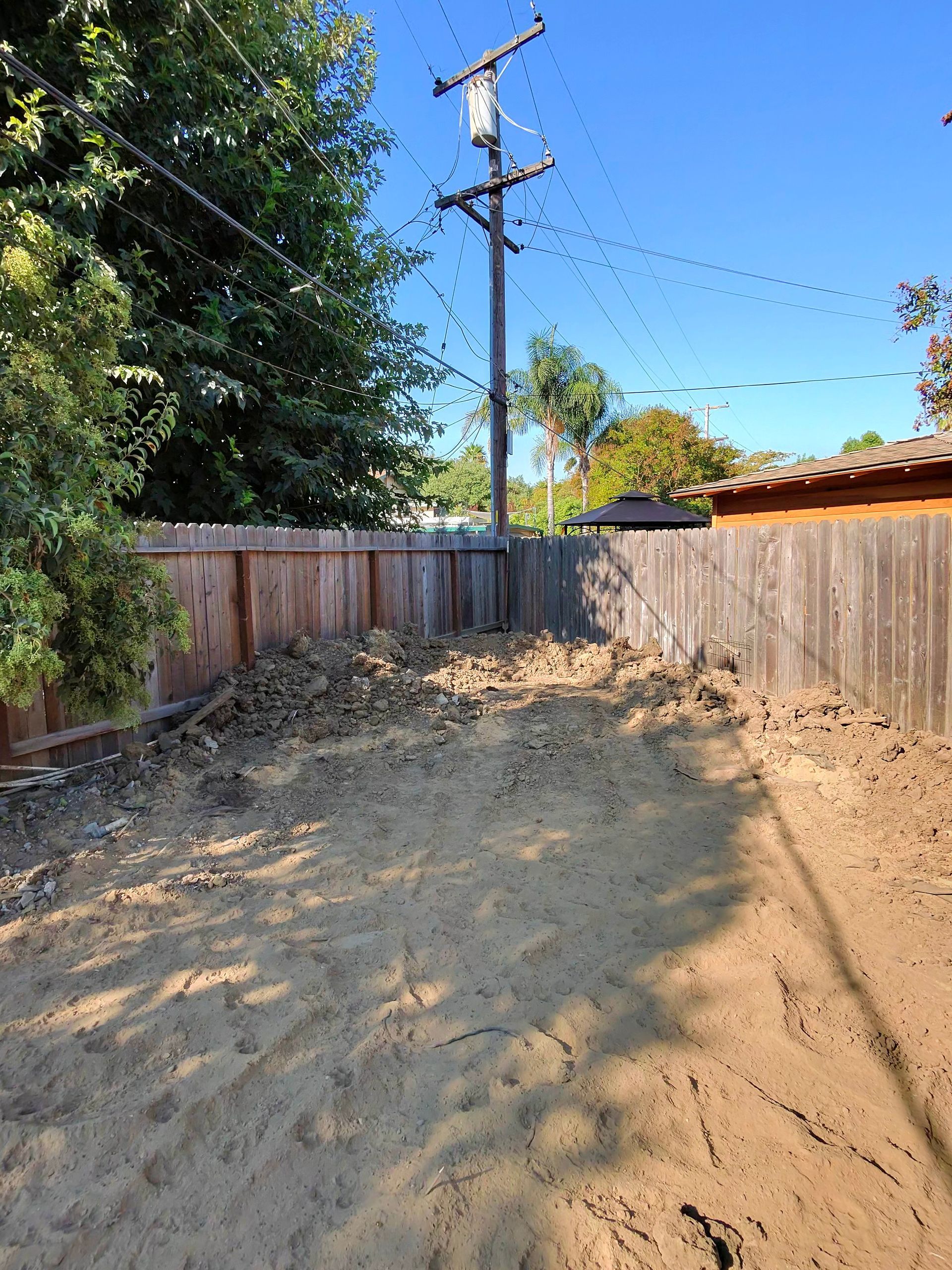 Dirt lot between wooden fences with a utility pole, trees, and a clear blue sky.