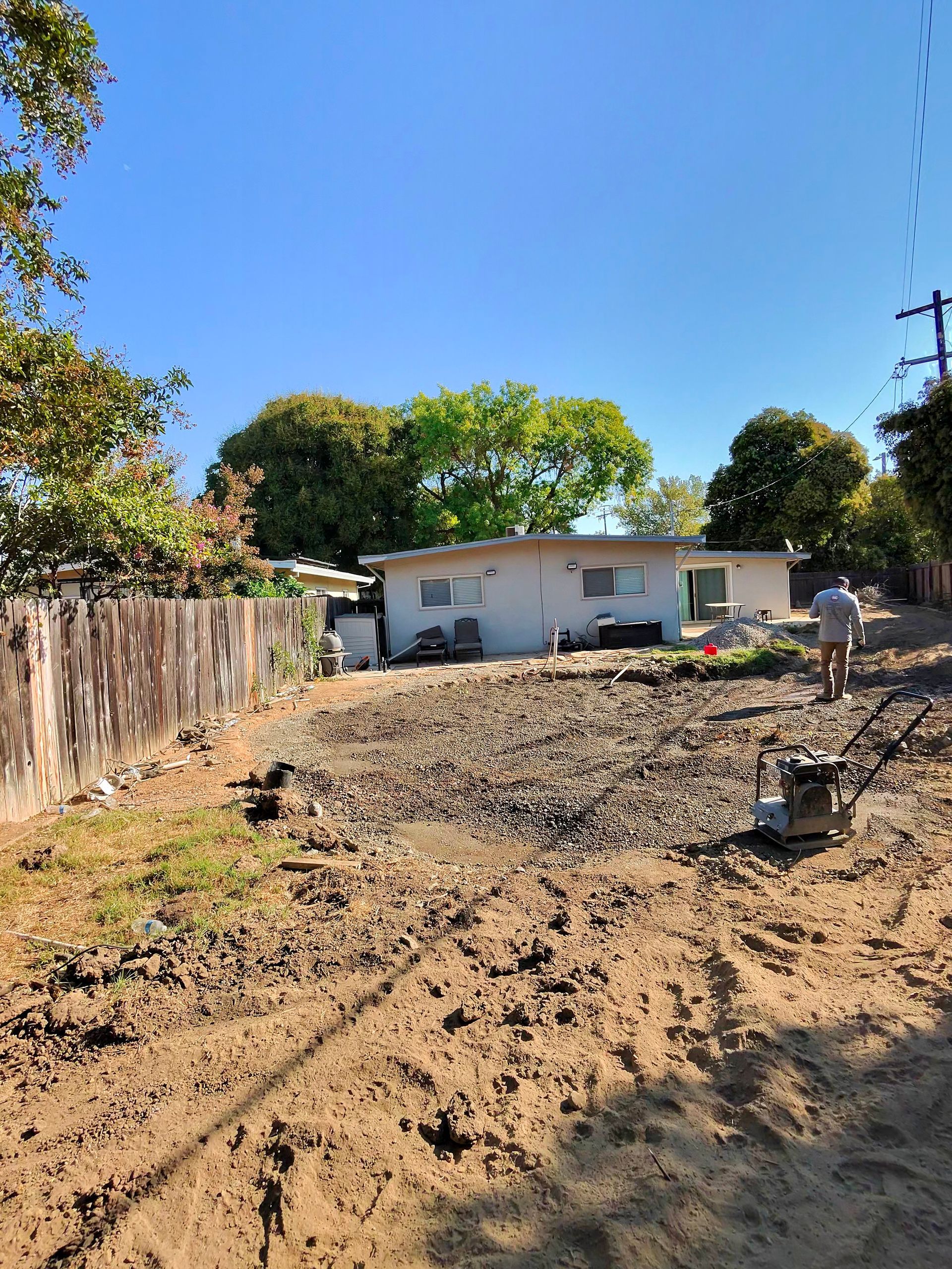 Backyard with dirt lot, small white house, wooden fence, and trees under a clear blue sky.