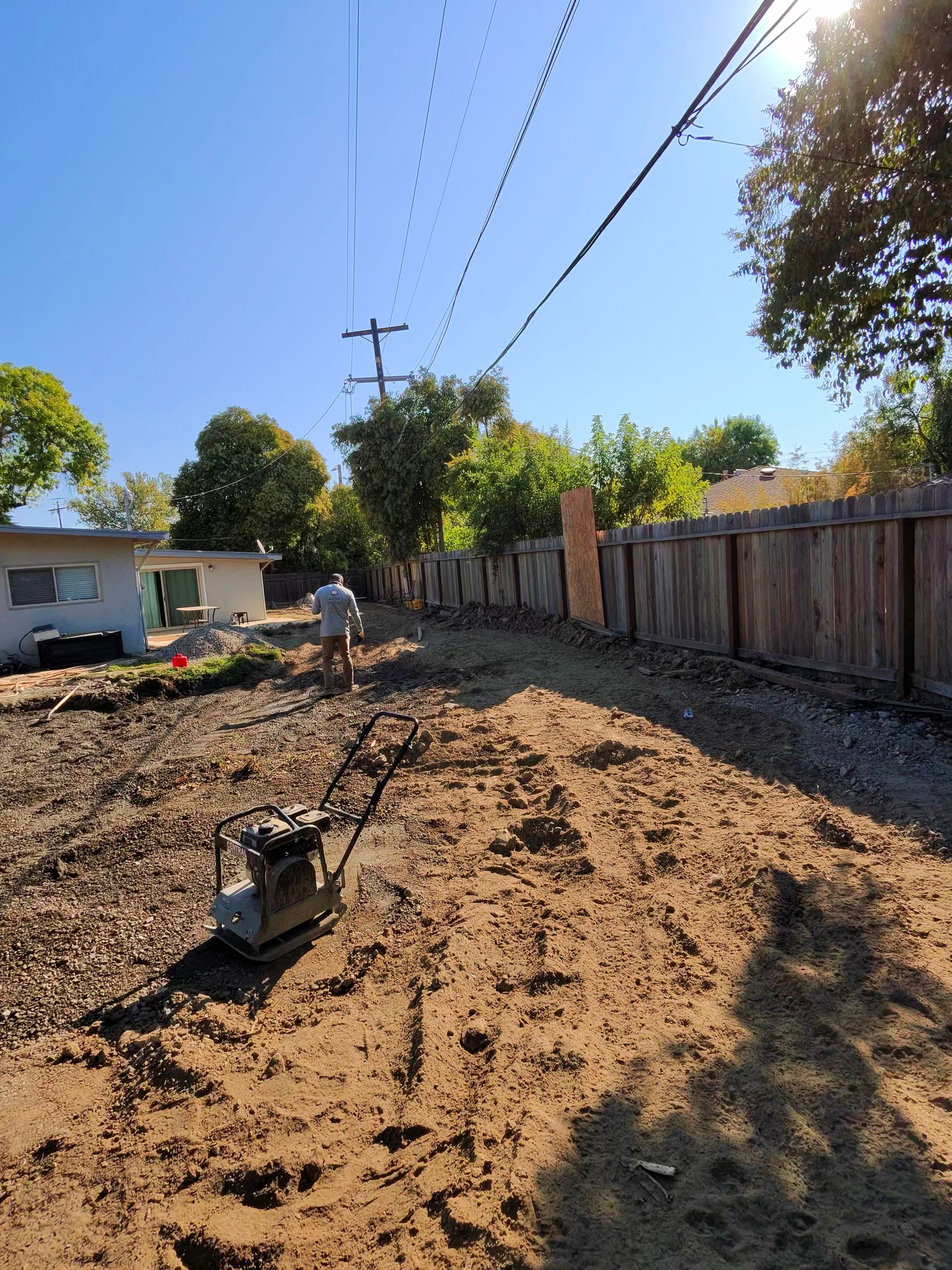 Dirt-covered yard with a wheelbarrow, fence, utility poles, and trees under a clear blue sky