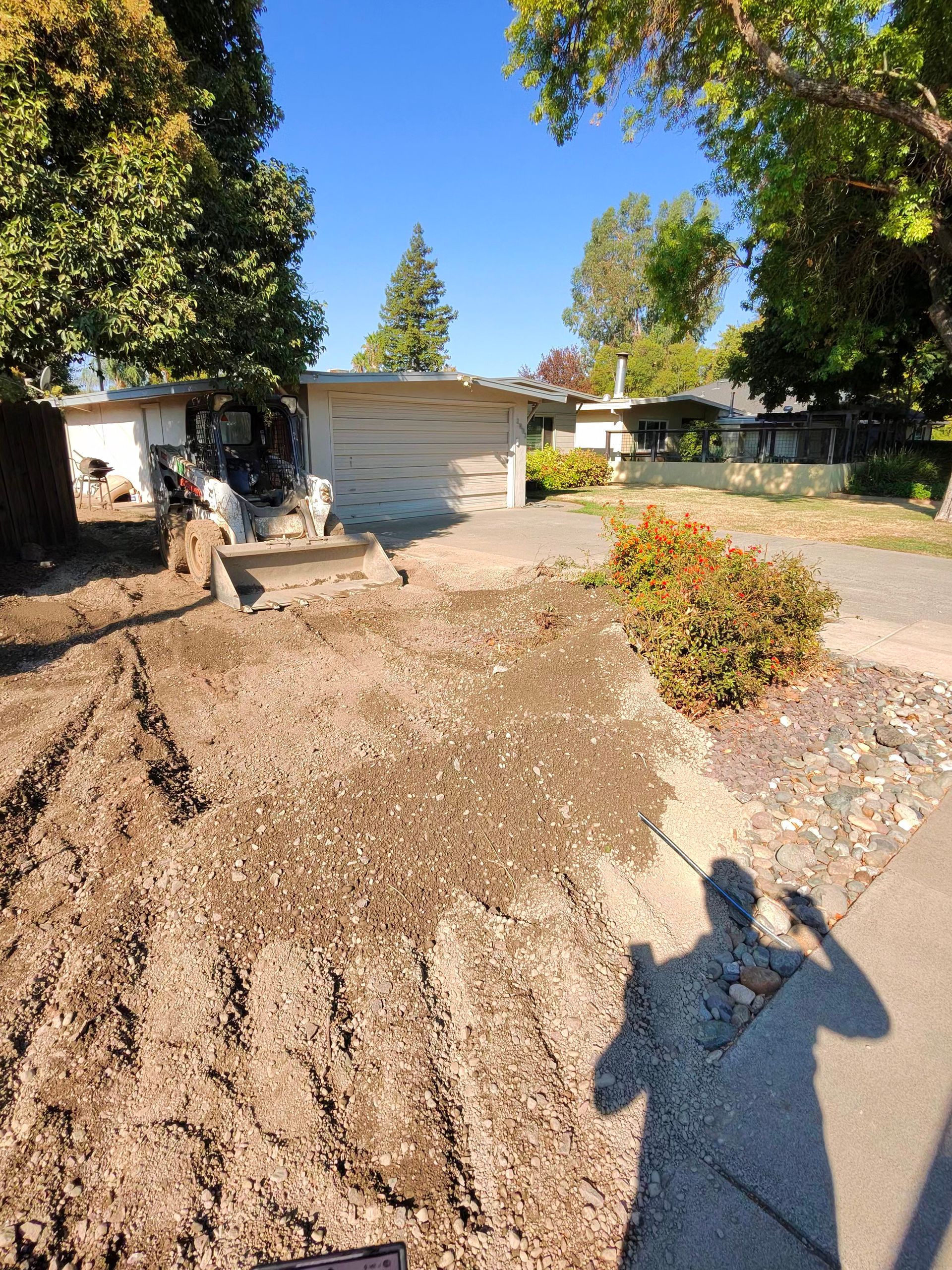 Sunlit gravel driveway with a white garage, trees, shrubs, and a person’s shadow in the foreground
