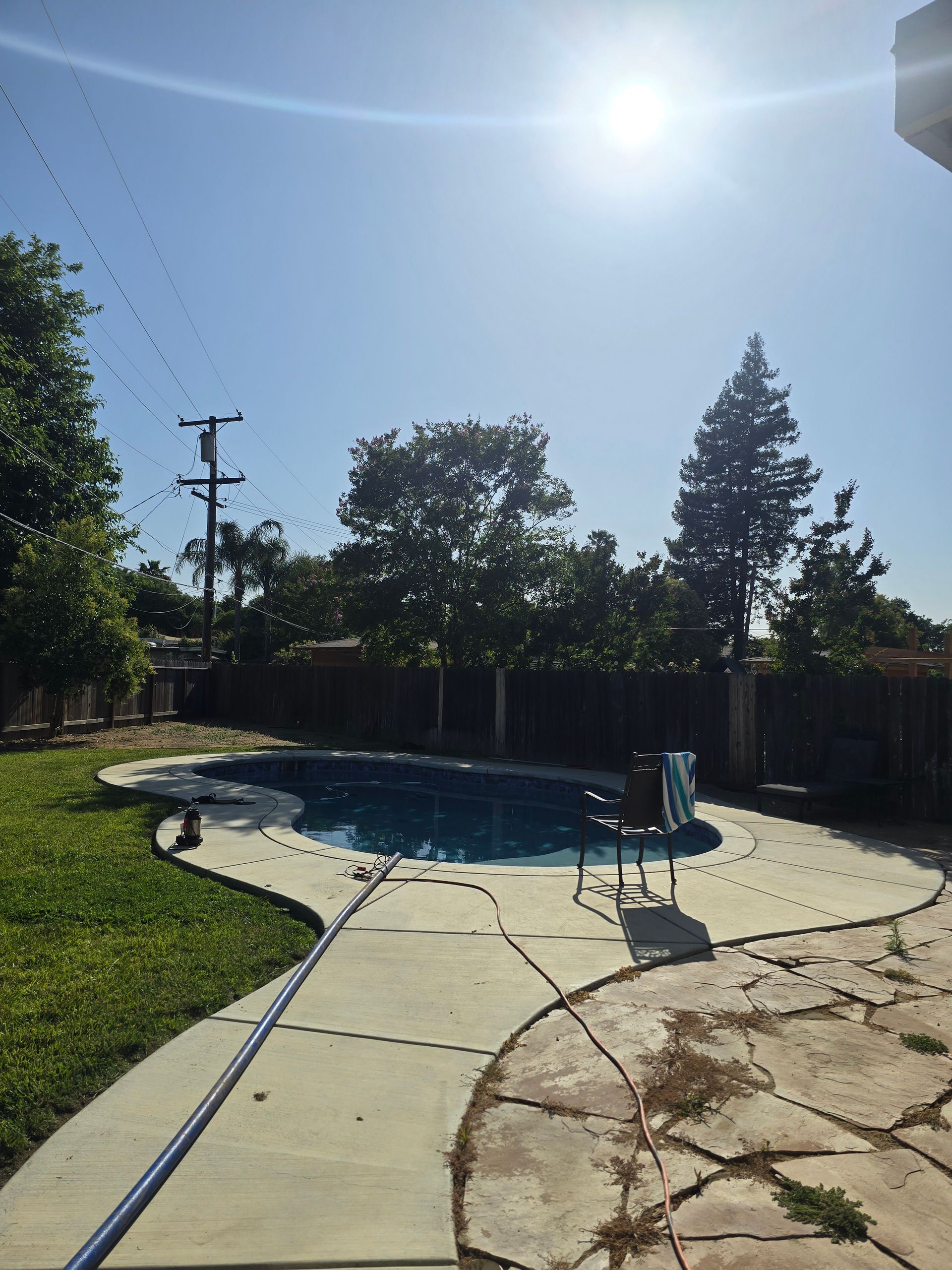 Backyard pool with cracked concrete patio, bright sun, and a hose stretched across the ground.