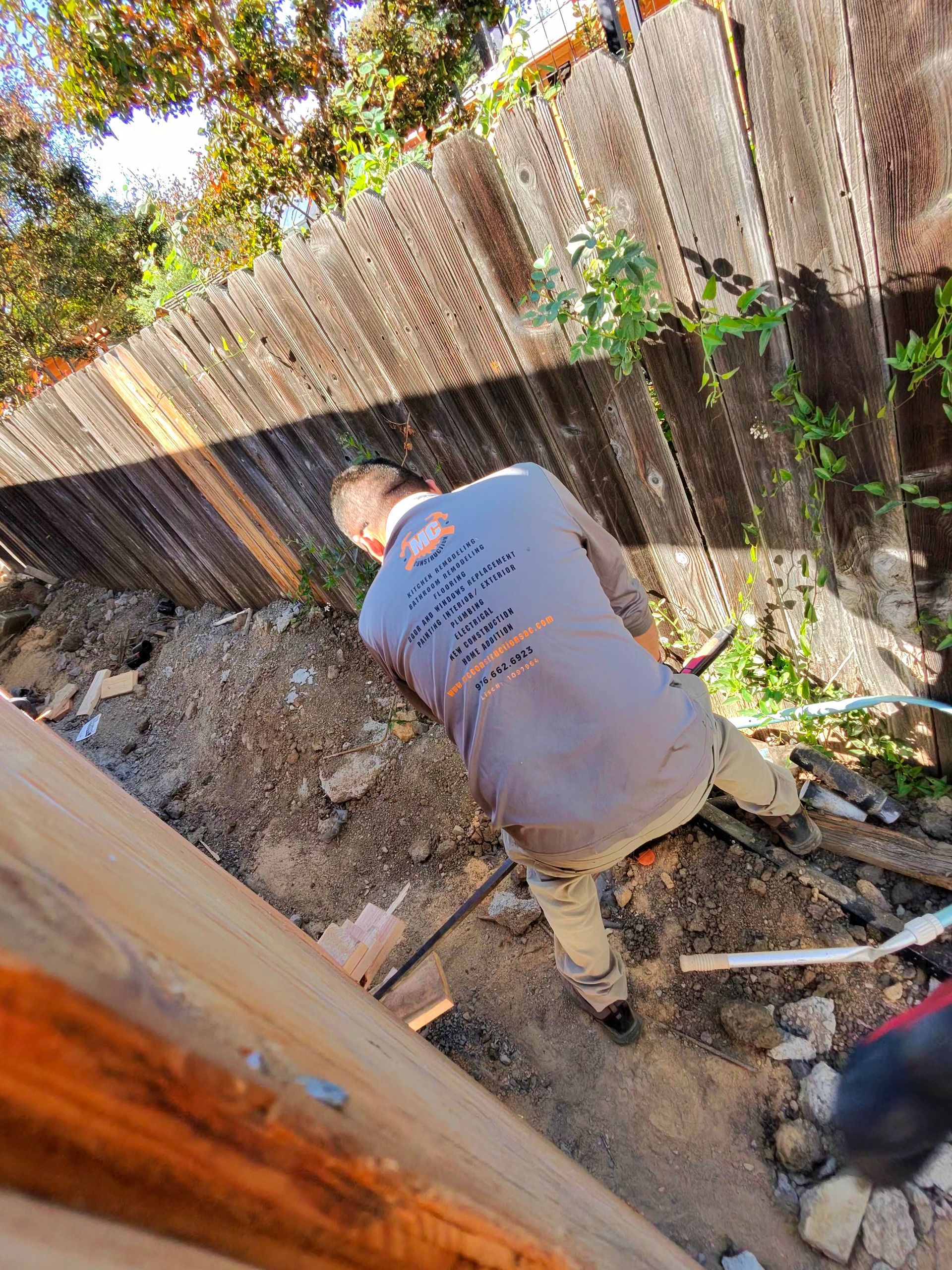 Person crouching in a dirt yard beside a weathered wooden fence, working near a hole and debris.
