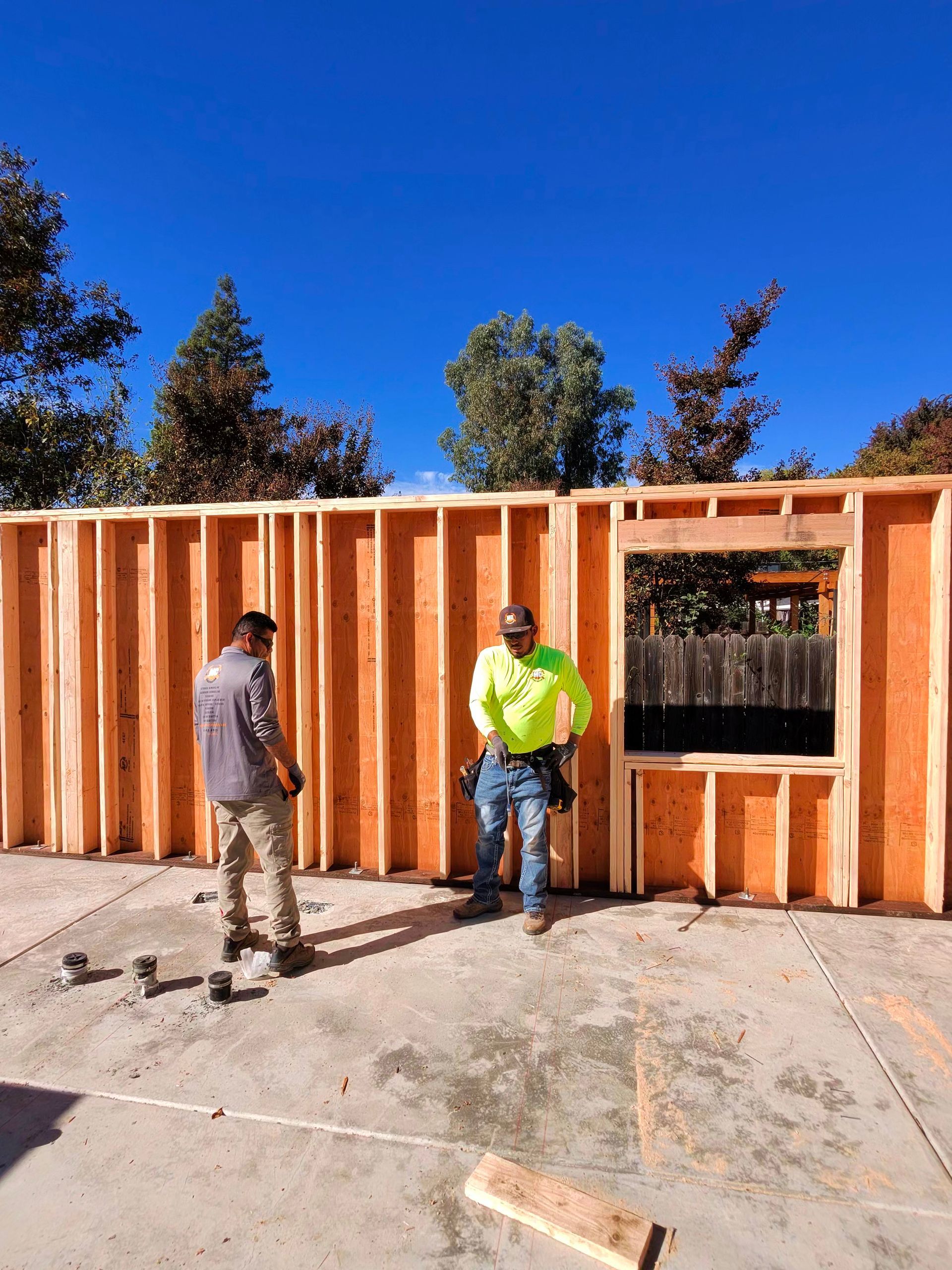 Two workers stand beside a partially framed wooden wall at an outdoor construction site.