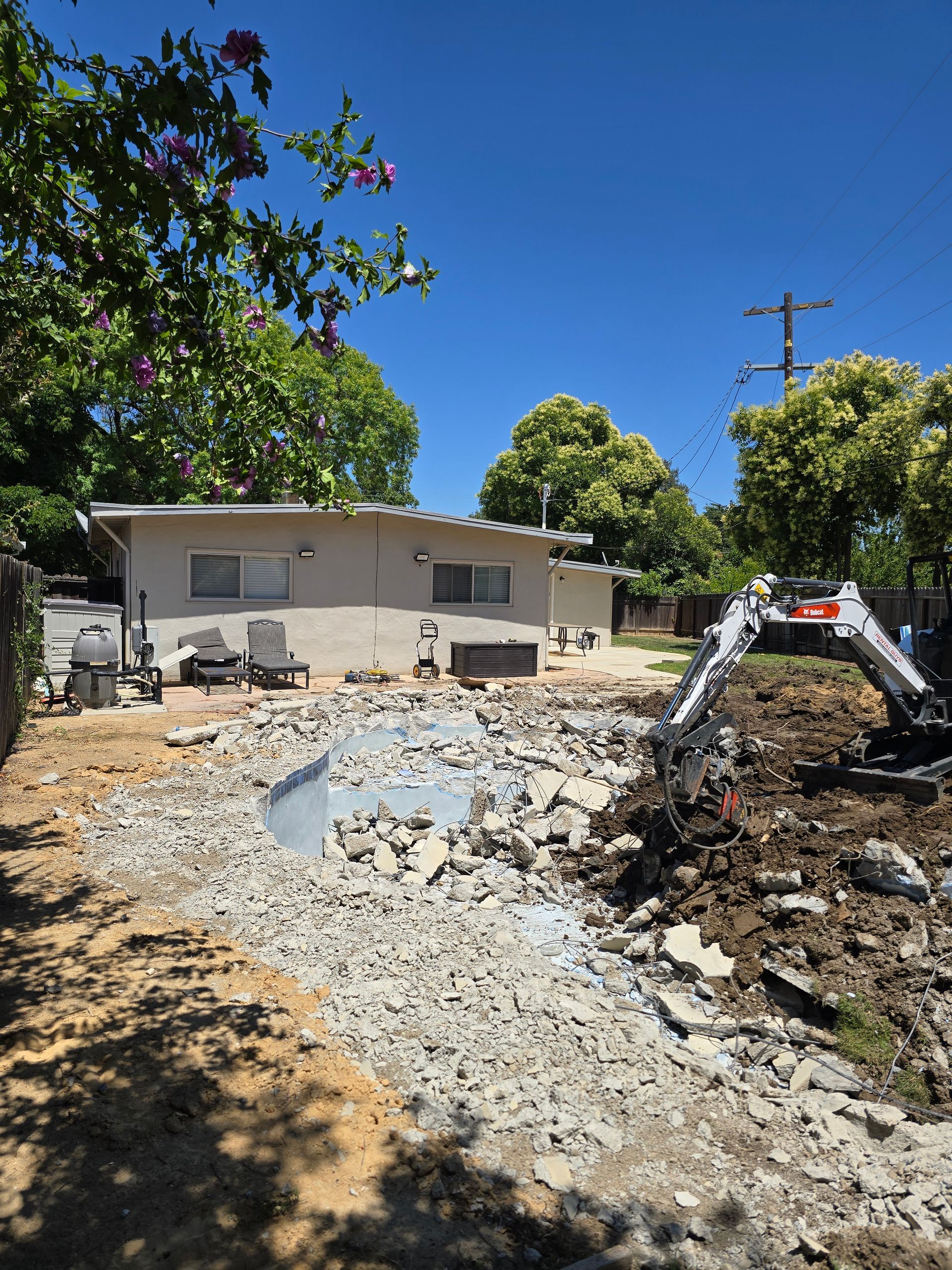 Backyard excavation beside a beige house, with gravel trench and an excavator arm on the right.