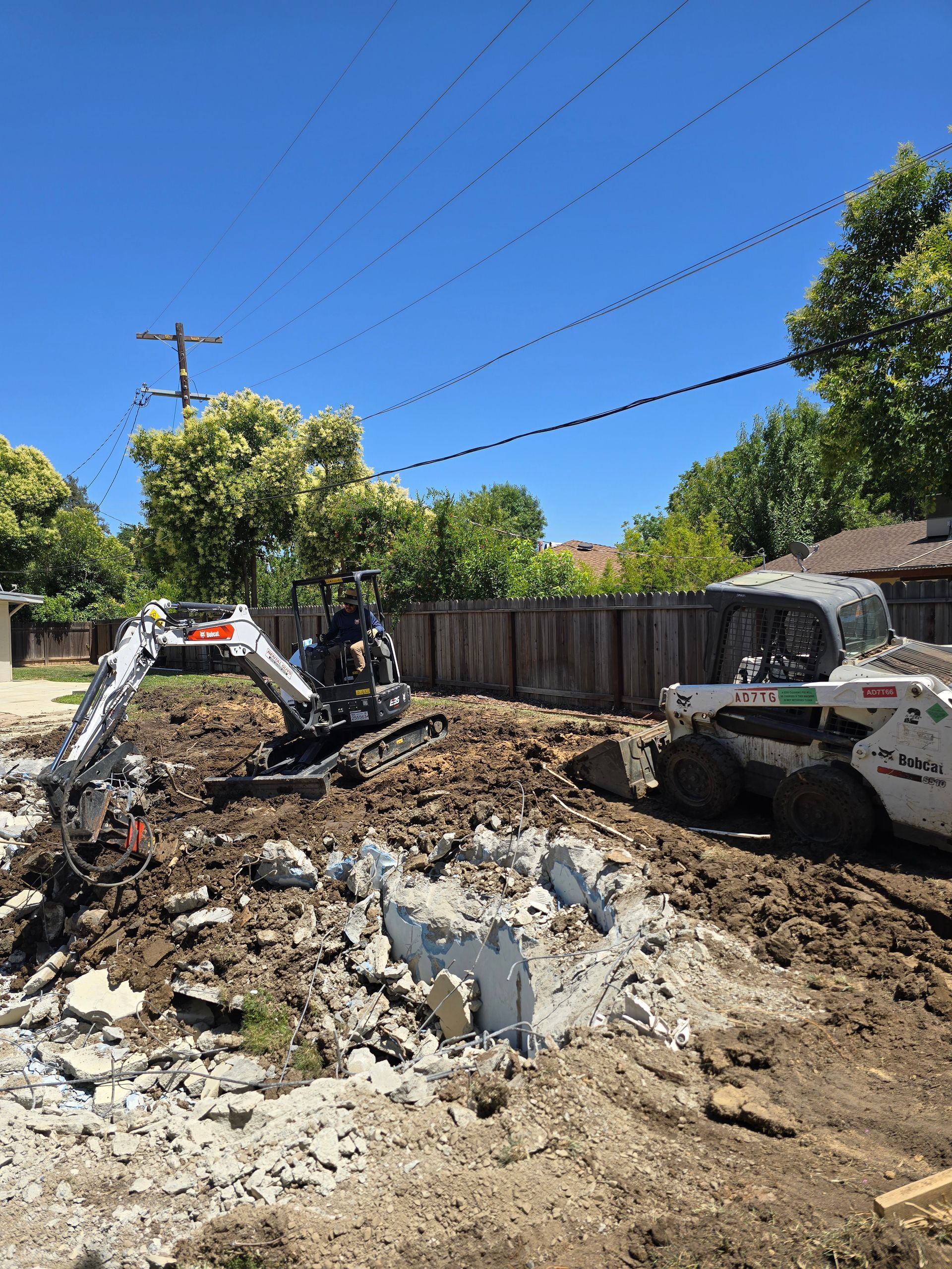 Demolition site with excavator digging rubble beside a partially demolished house on a sunny day