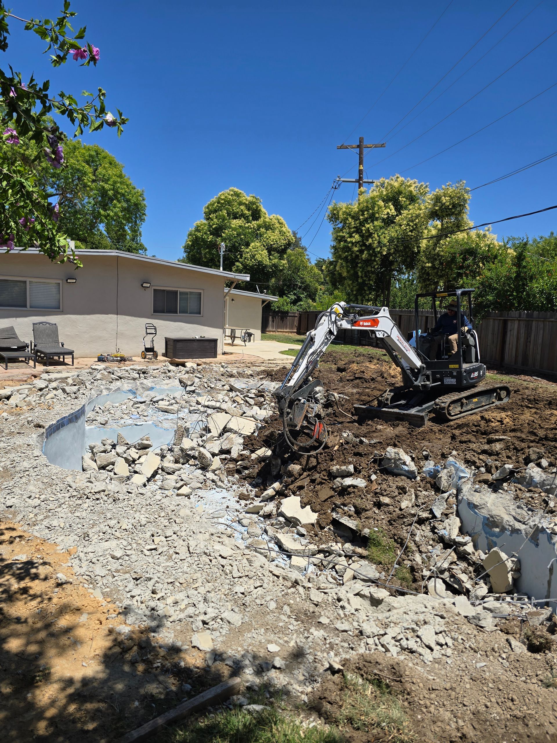 Excavator digging a trench beside a house under a bright blue sky, with gravel and dirt piled nearby.