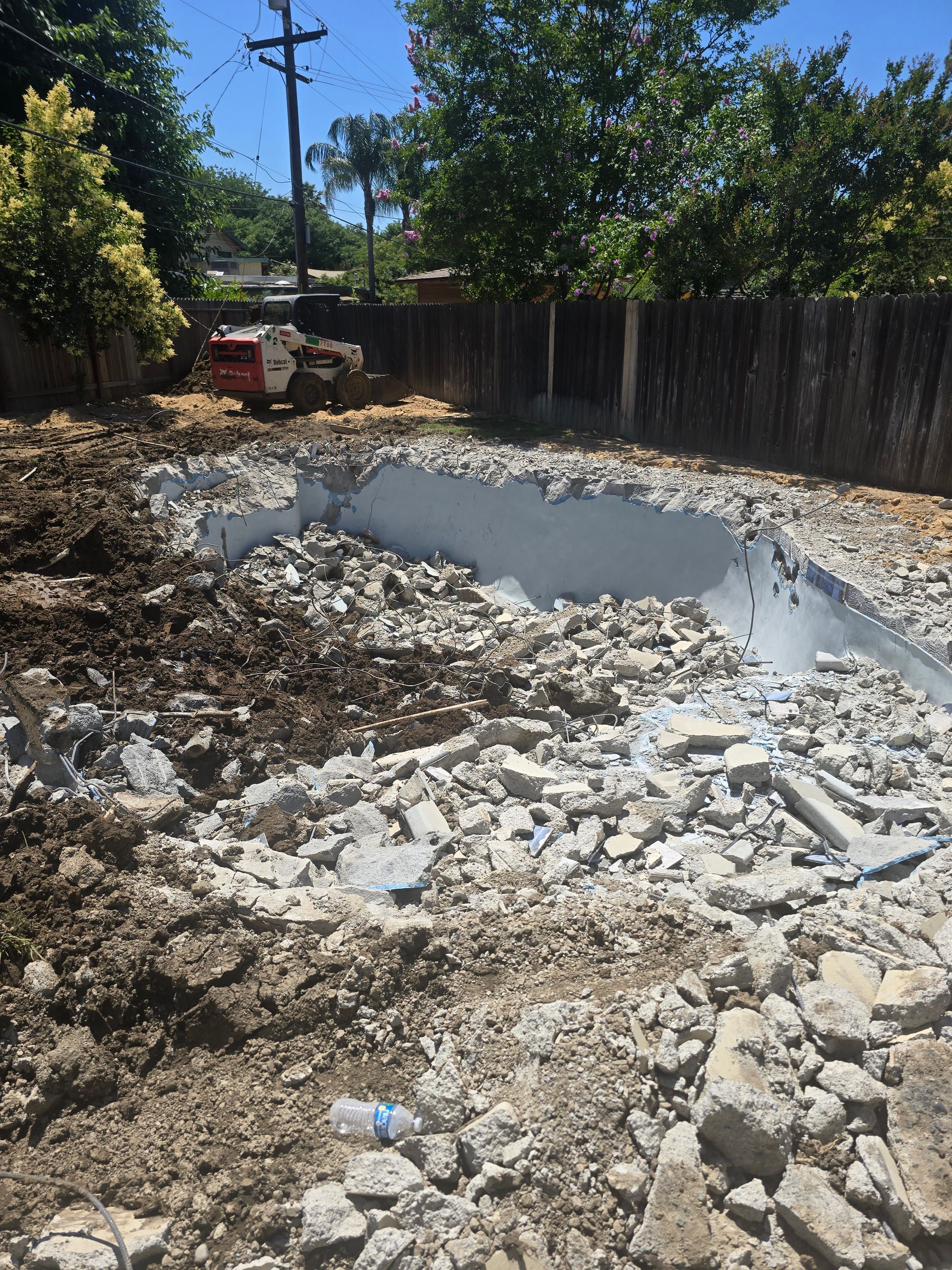 Demolished concrete foundation with rubble, dirt, and a wooden fence under trees in a sunny yard.