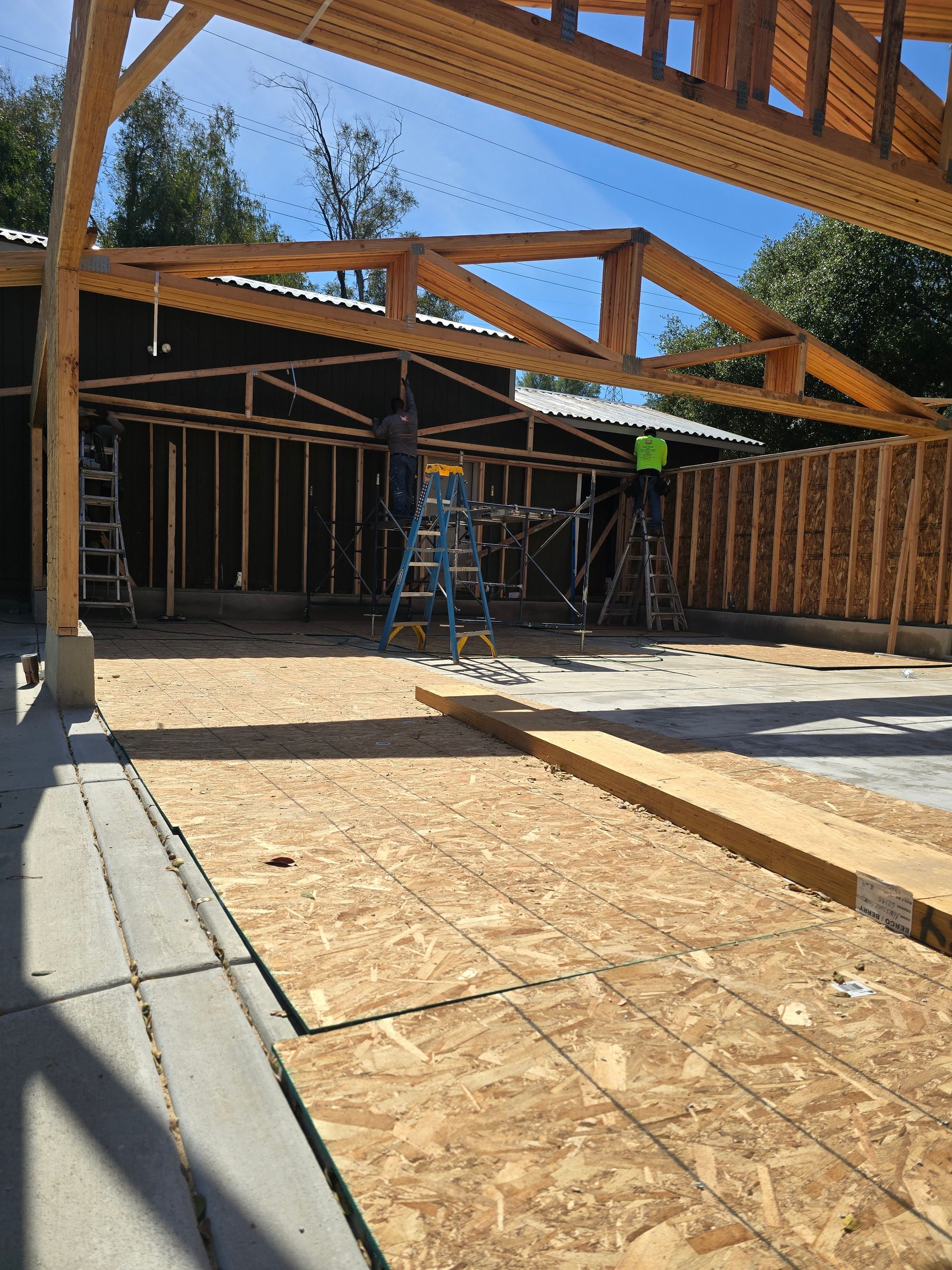Partially framed house under construction with roof trusses, plywood floor, and scaffolding in bright sunlight