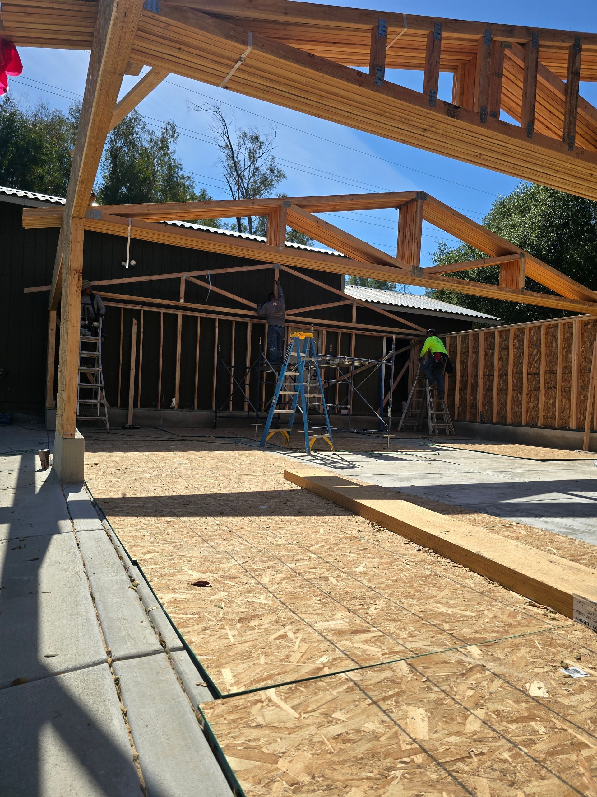 Partially built wooden house interior with exposed trusses, scaffolding, and workers on a concrete slab