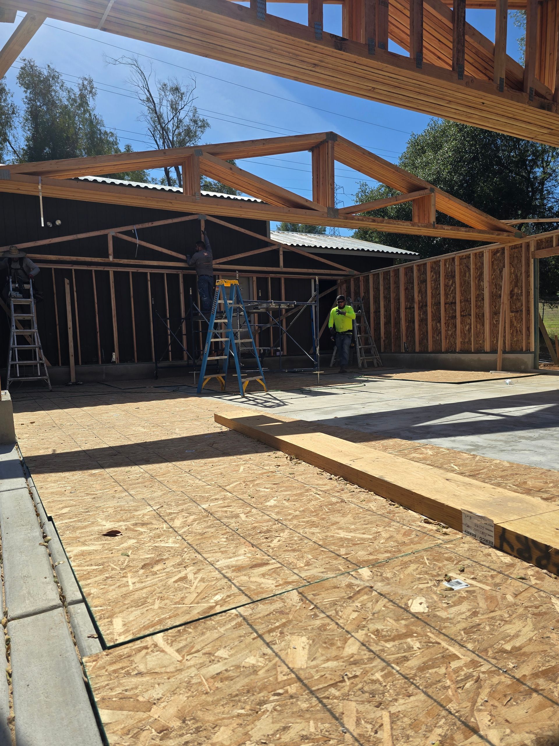Construction site with exposed wooden roof trusses, open framing, and workers under a bright blue sky