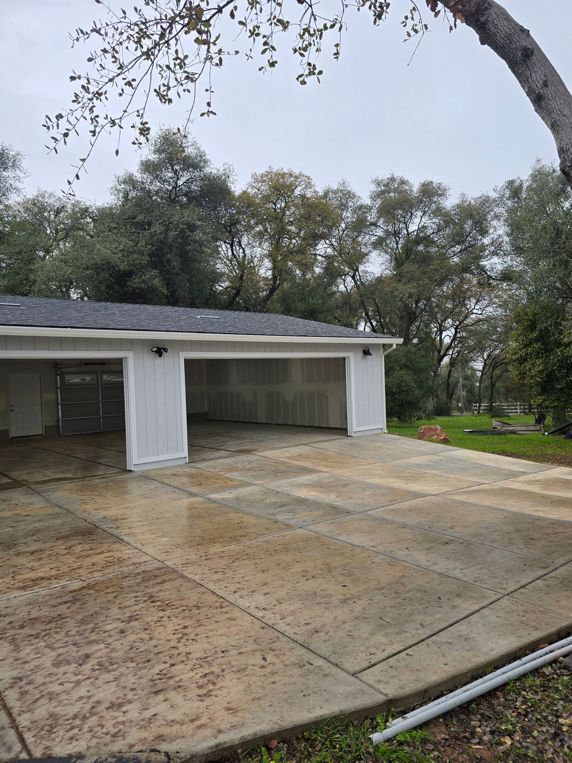 Rainy driveway beside open garage with wet concrete and trees in the background