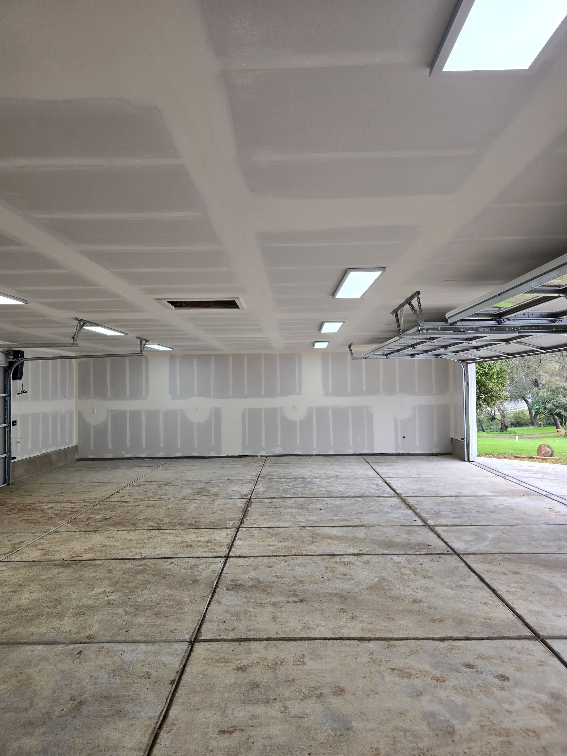 Empty unfinished garage interior with exposed drywall and open door to a green yard