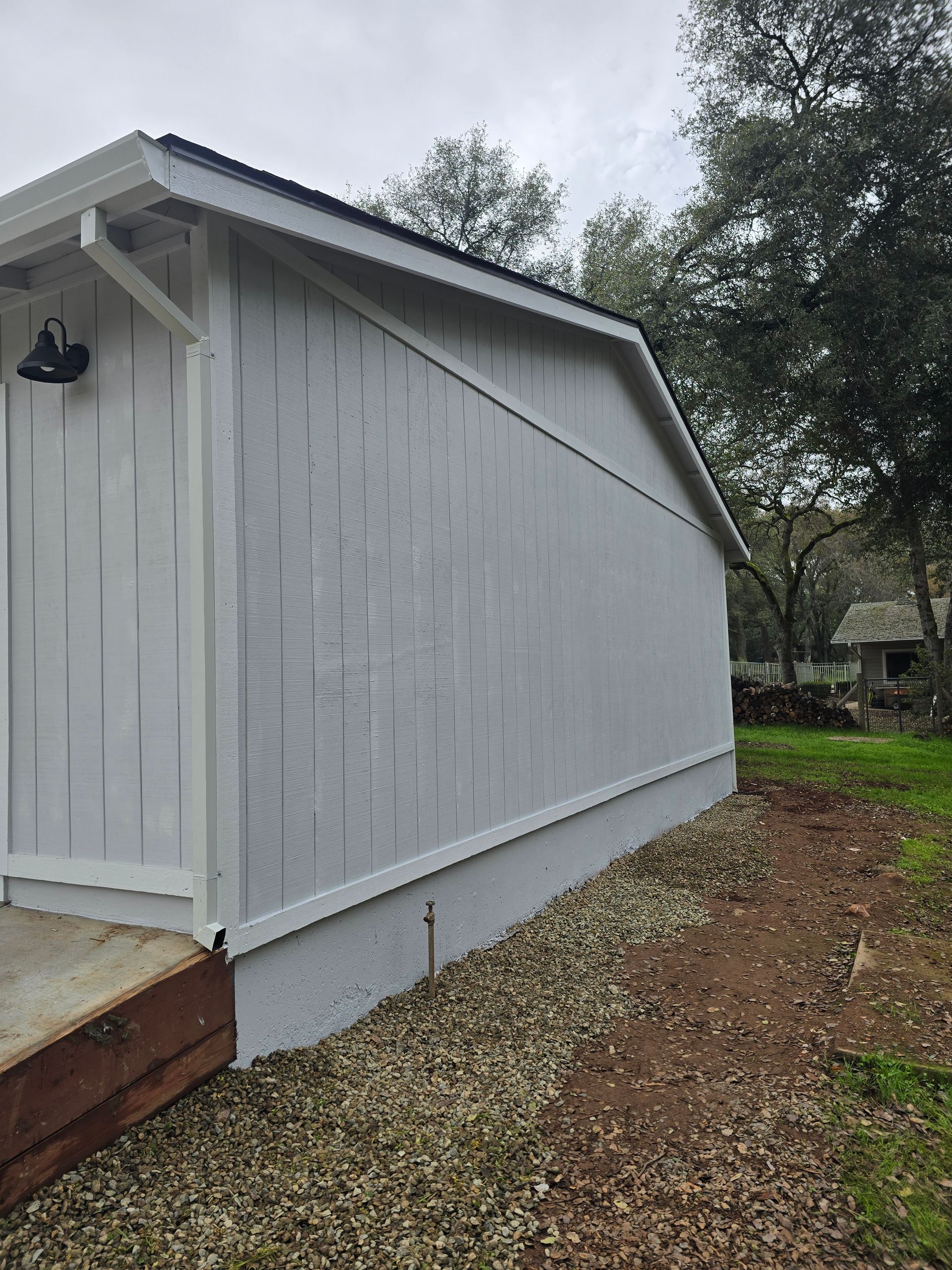 Gray house side with white trim and a small porch, set beside a gravel path and trees.