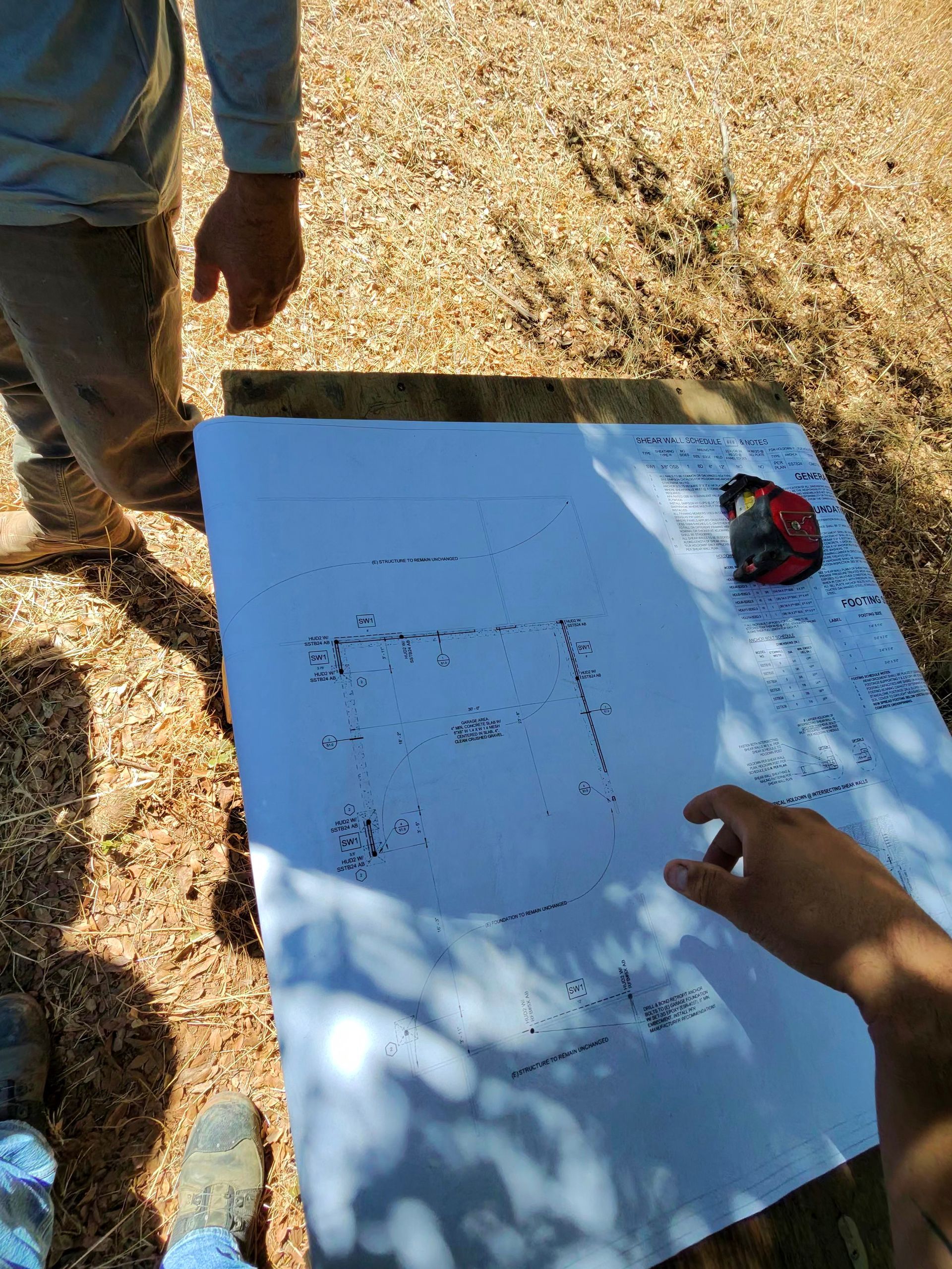 People examining a large white sheet with notes on sunlit gravel outdoors