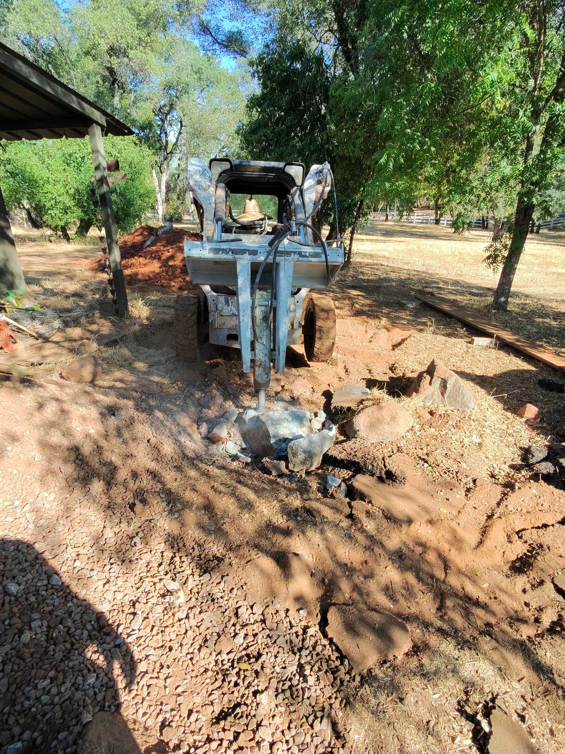 Small tractor digging a muddy trench in a wooded yard with scattered leaves and trees.
